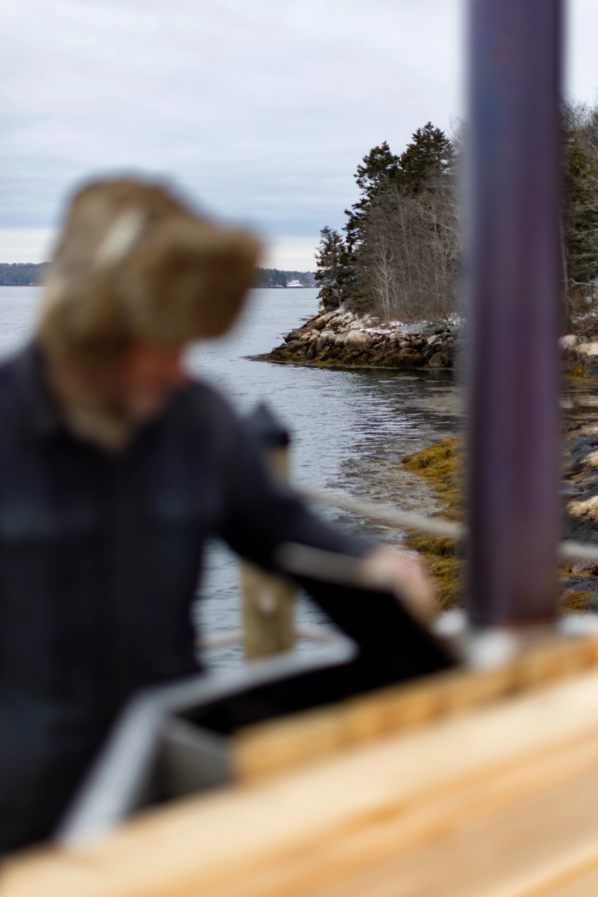 A person wearing a hooded jacket and a knit hat, standing on a dock near a body of water with trees in the background, blurred in the foreground.