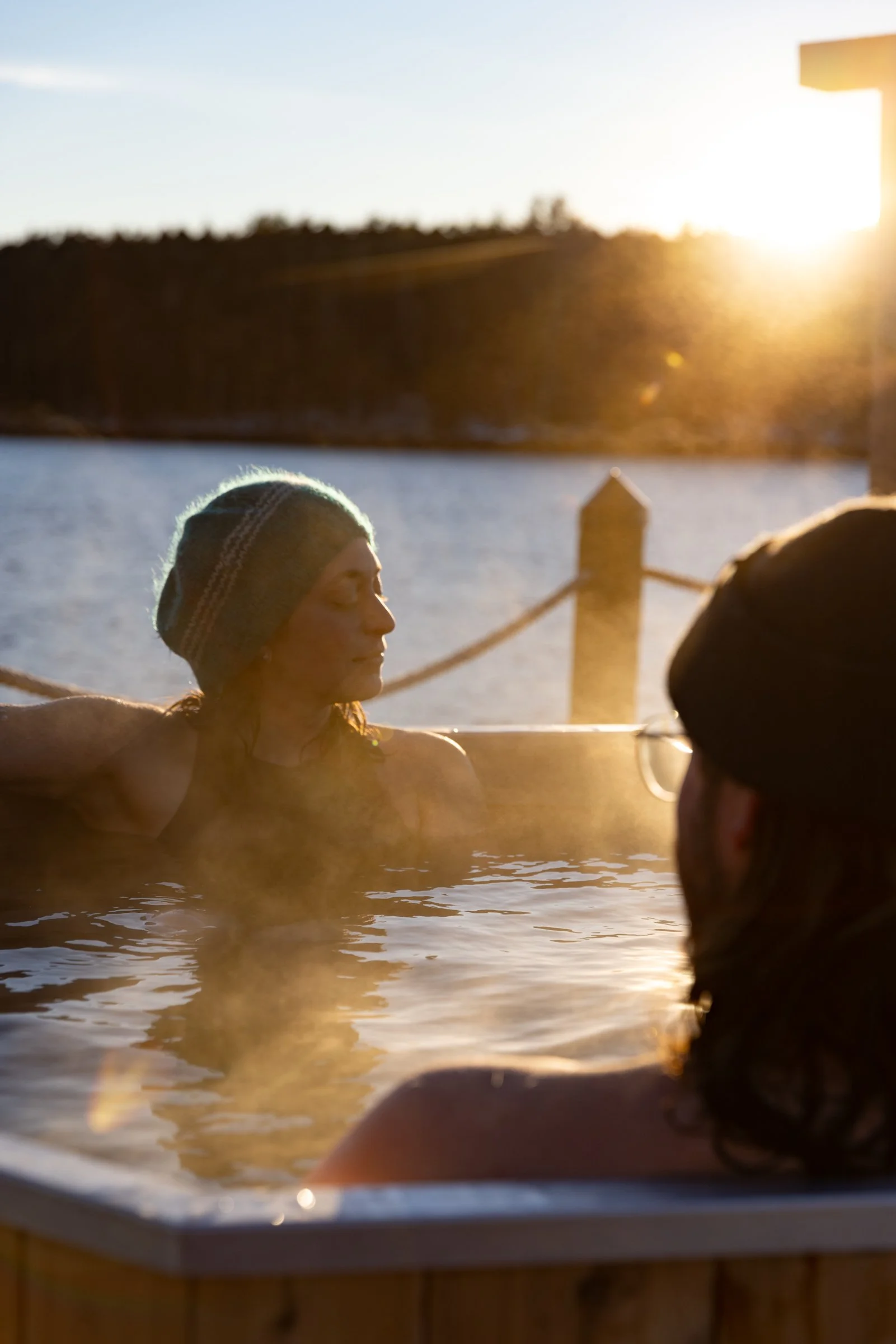 Two people relaxing in a hot tub outdoors during sunset, with a body of water and a distant shoreline in the background.