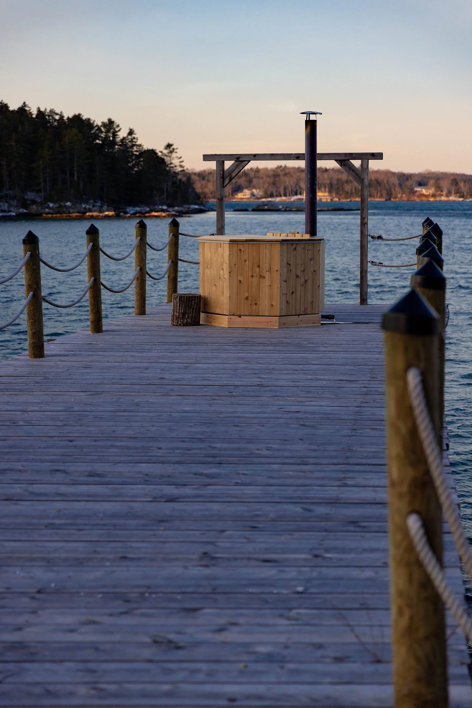 A wooden dock extending over a body of water with a small hot tub and a pipe chimney at the end, surrounded by trees and a distant shoreline.