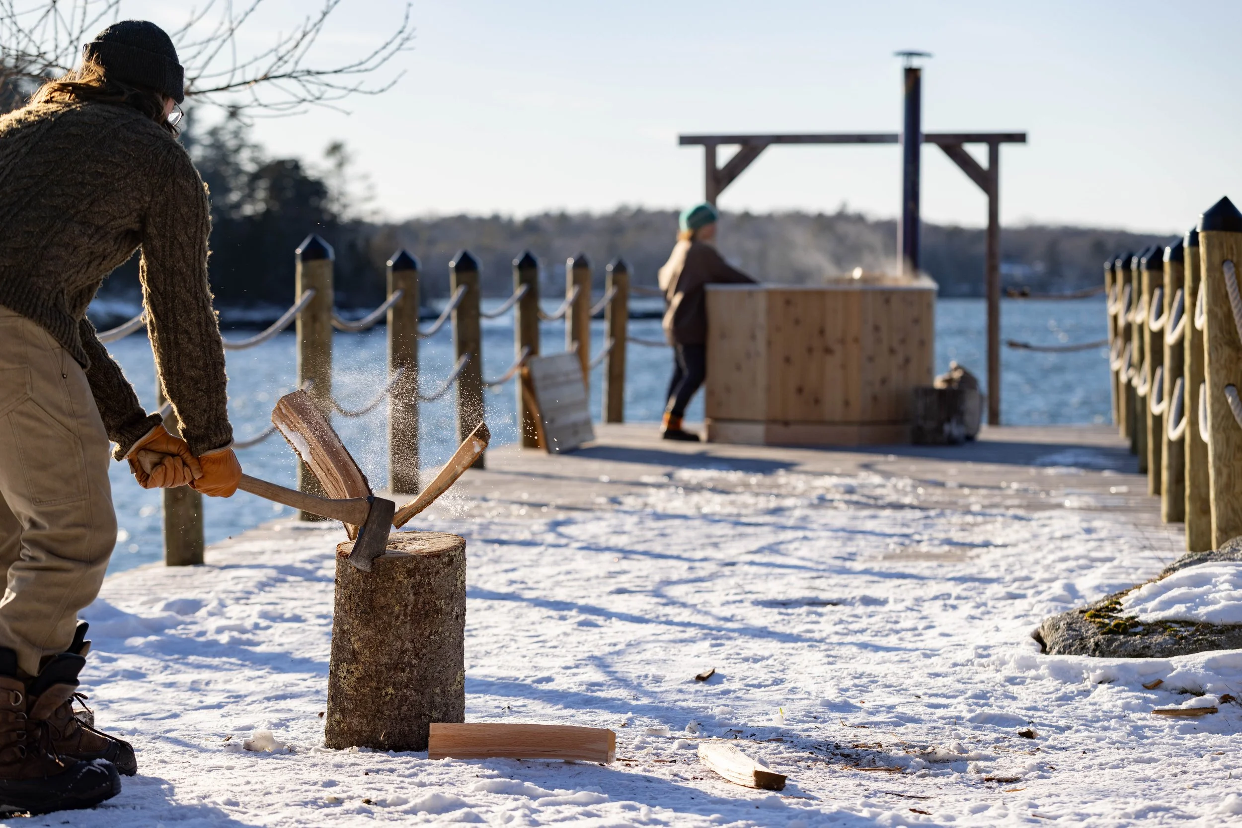 Person chopping wood with an axe on a snowy dock by the water, with two people in the background near a wood fired aluminum hot tub.
