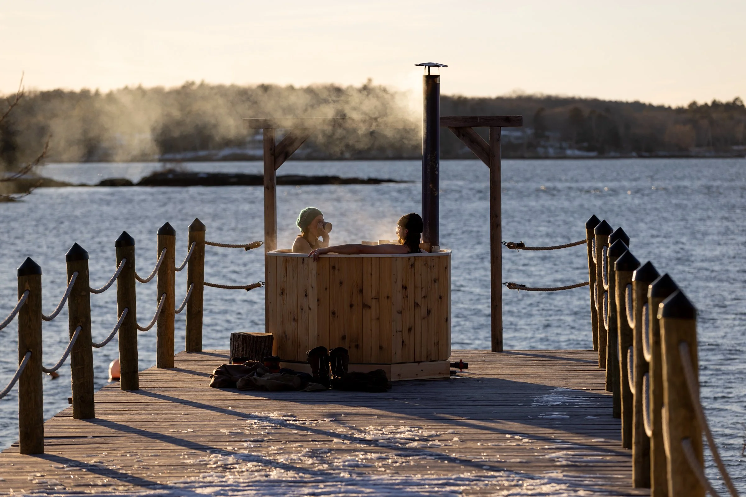 Two people relaxing in a wood fired aluminum hot tub on a dock by the ocean during winter with snow, steam rising from the hot water, and trees in the background.