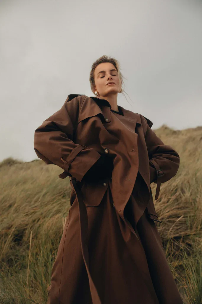 A woman standing outdoors against a rocky landscape and cloudy sky, dressed in a beige oversized coat and gloves, looking off into the distance.