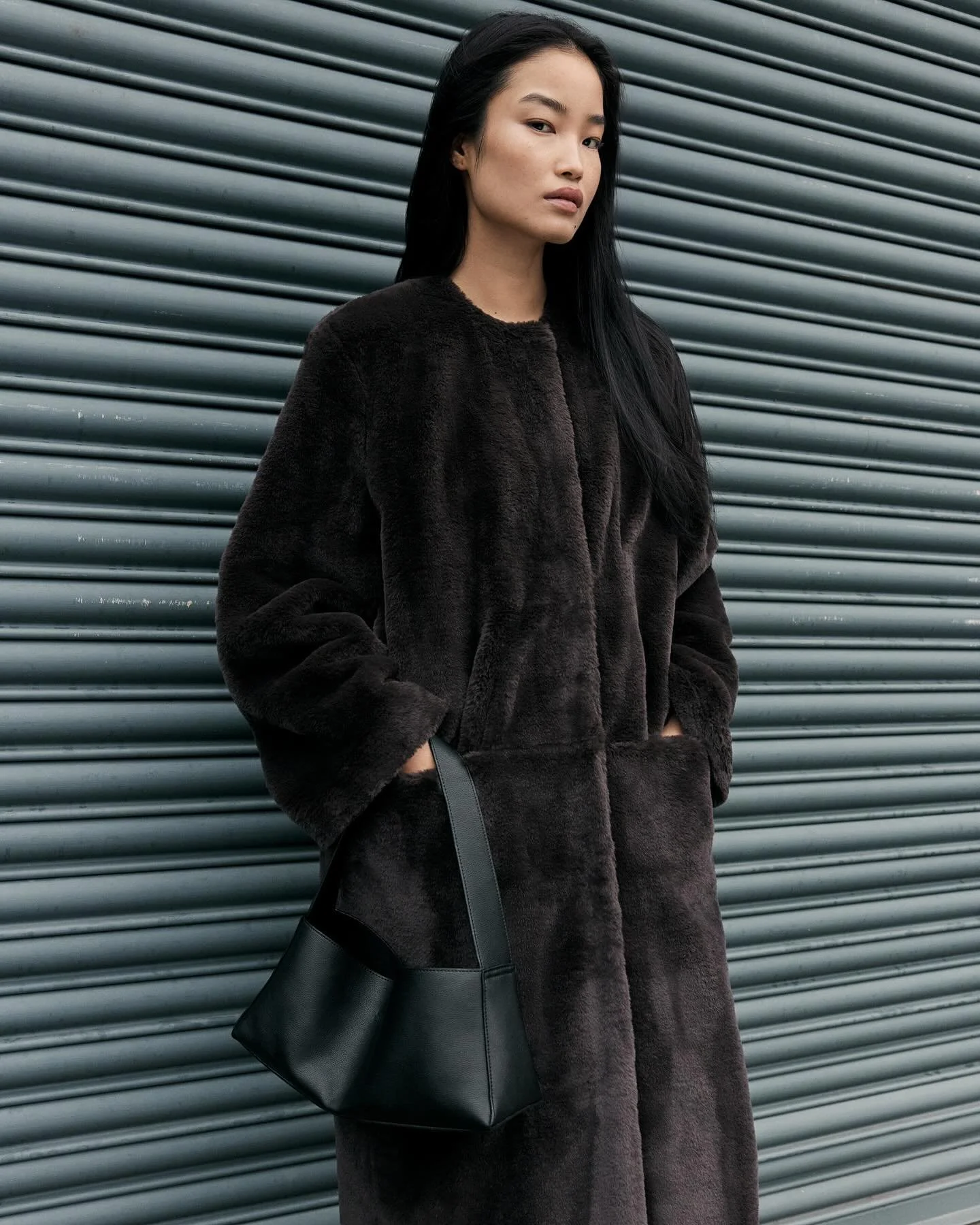 A woman with long black hair wearing a dark, plush coat and carrying a black leather handbag stands against a metallic, horizontal-slatted background.