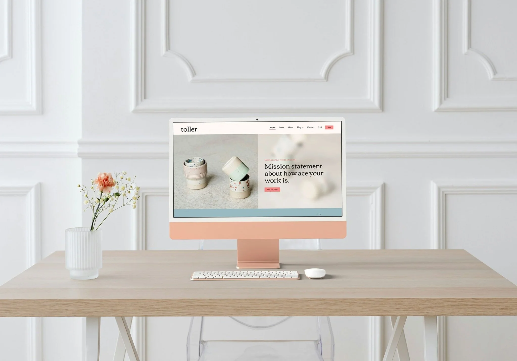 A minimalist workspace with a wooden desk, a desktop computer displaying a website, a white keyboard, a white mouse, and a vase of pink flowers set against white paneled walls.