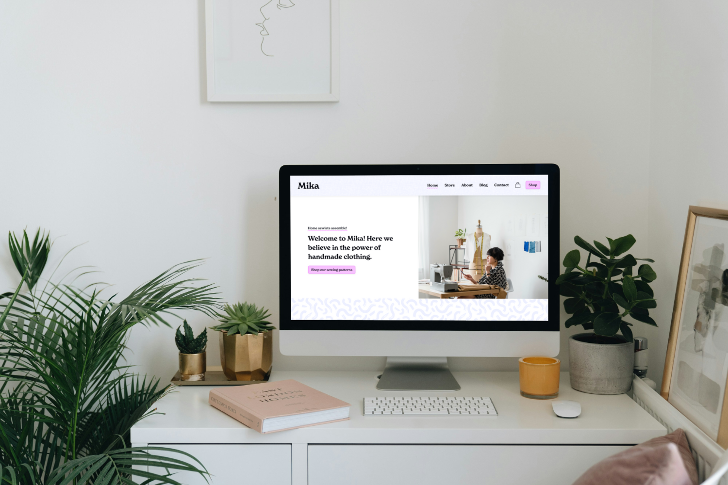 A computer monitor displaying a website for a handmade clothing store called Mika, set on a white desk surrounded by plants, a book, and a framed picture.