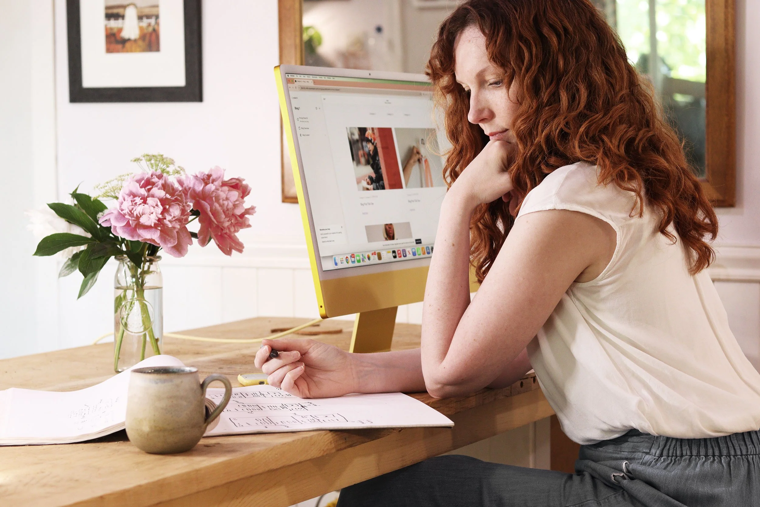 A woman with curly red hair sitting at a desk, looking at a computer monitor. There is a notebook, a mug, and a vase with pink flowers on the desk. The room has a window and framed pictures on the wall.