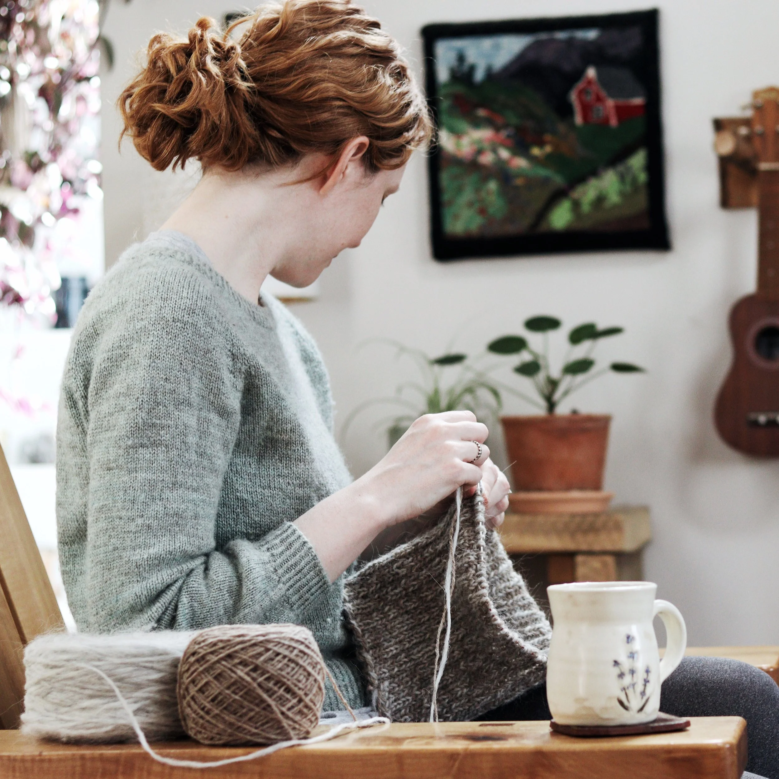 A woman knitting with yarn balls on a wooden table, a potted plant and painting in the background.
