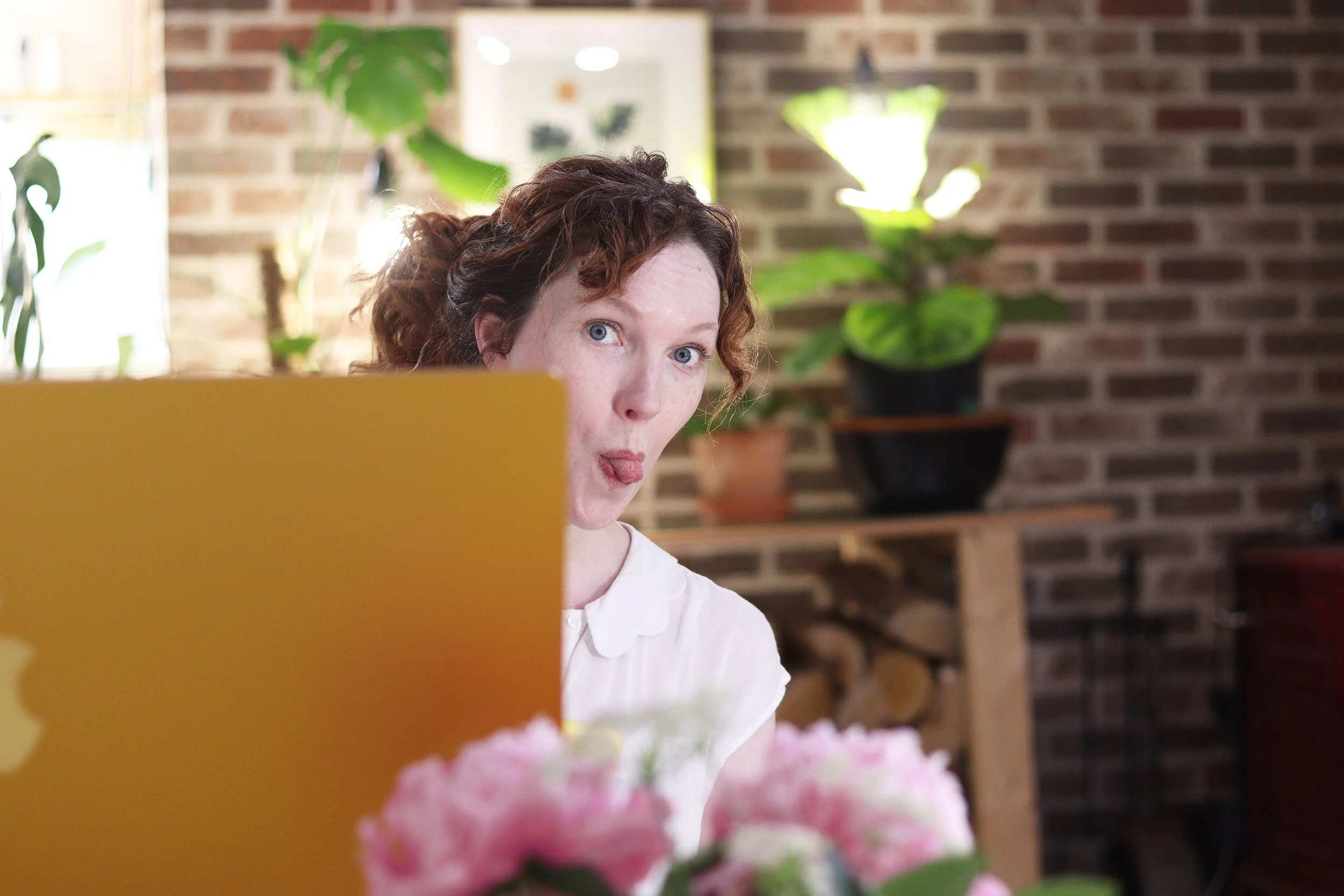 A woman with curly red hair making a funny face at her laptop, which is partly visible in the foreground. The background features a brick wall, green plants, and a window letting in natural light.