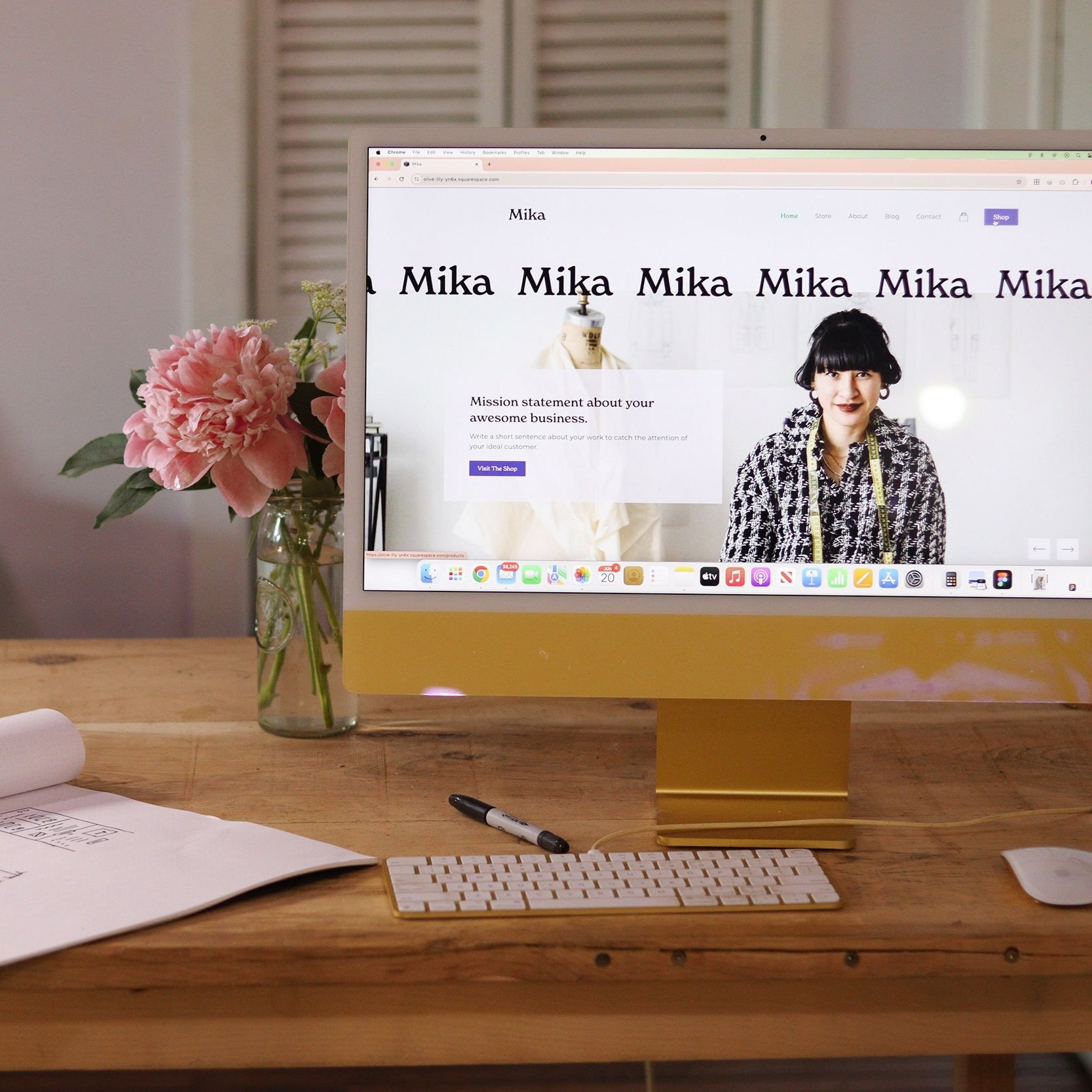An Apple desktop computer on a wooden desk displays a webpage with the name Mika, a woman wearing a patterned top, and a message about an awesome business. Next to the monitor is a vase with pink flowers, a black marker, a white keyboard, and a white