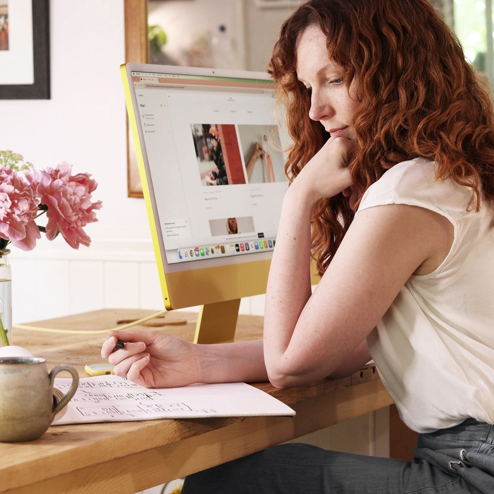 A woman with curly red hair sitting at a wooden desk, working on a computer with a pink flower vase nearby.