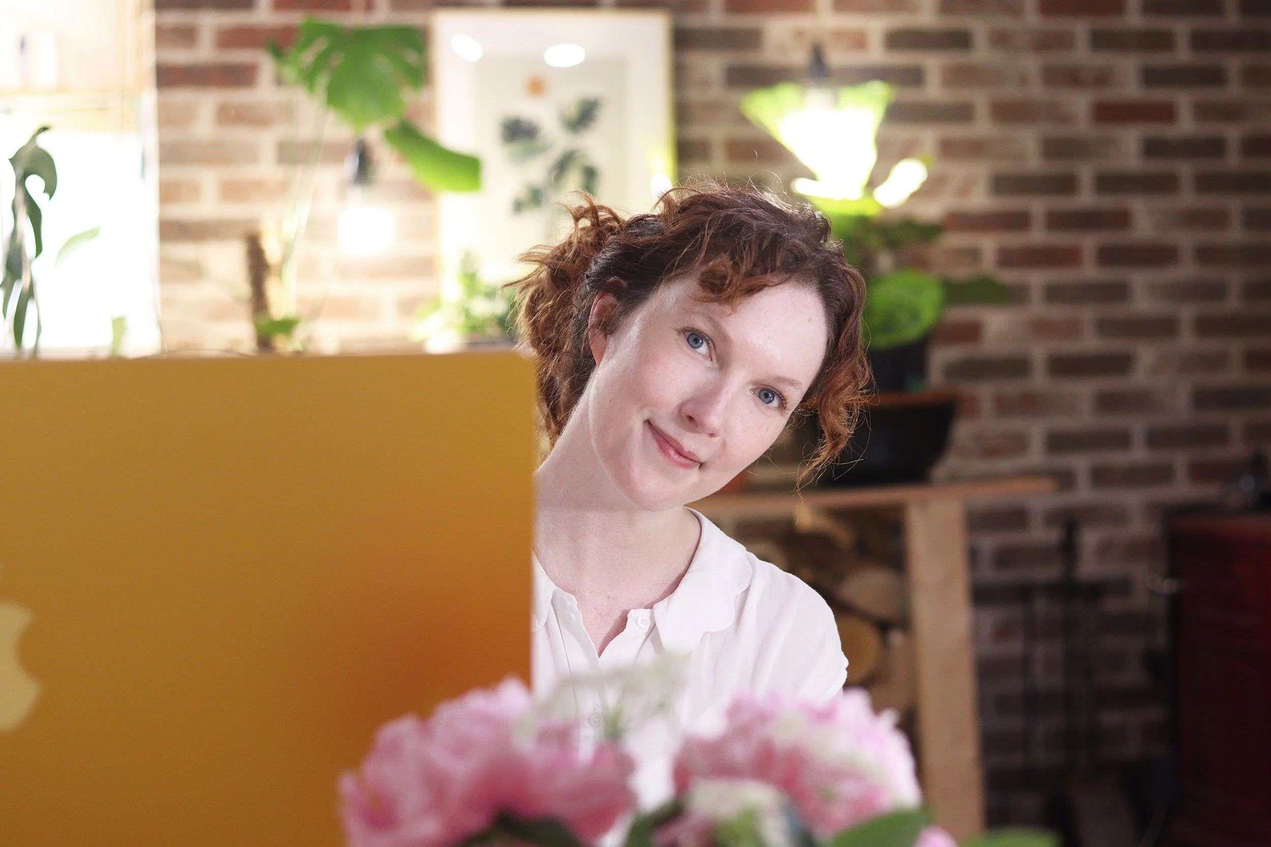A woman with curly red hair and blue eyes sitting in a cozy room, smiling at a yellow laptop with pink flowers in foreground, greenery and brick wall in background.
