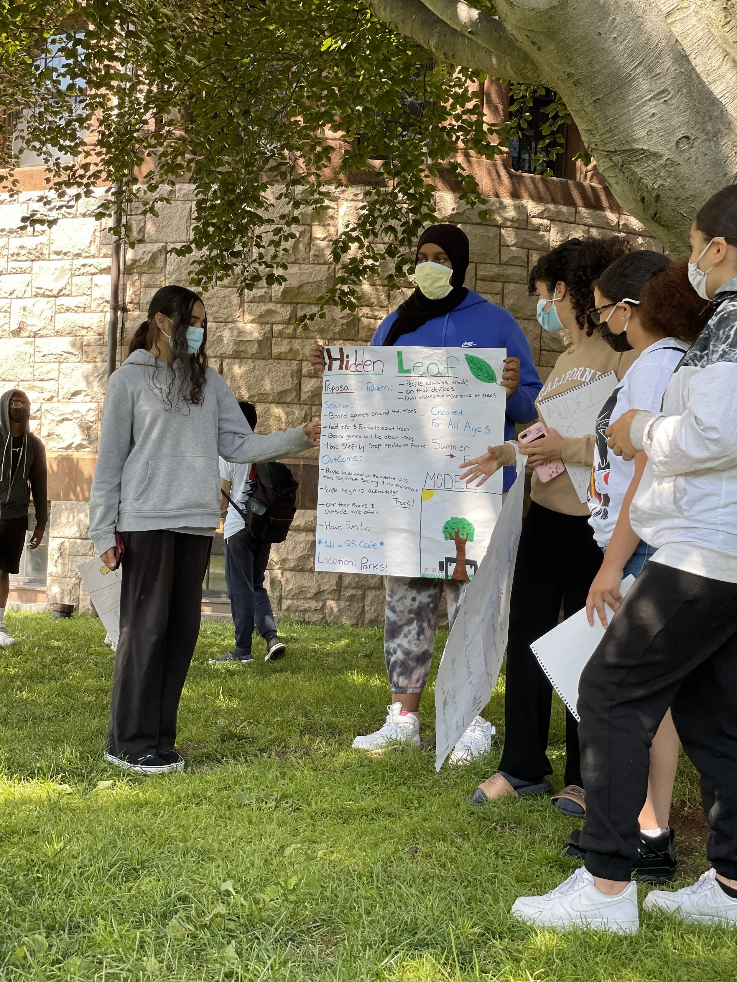 Group of kids outdoors near a stone building and tree, wearing masks, holding a poster about environmental awareness.