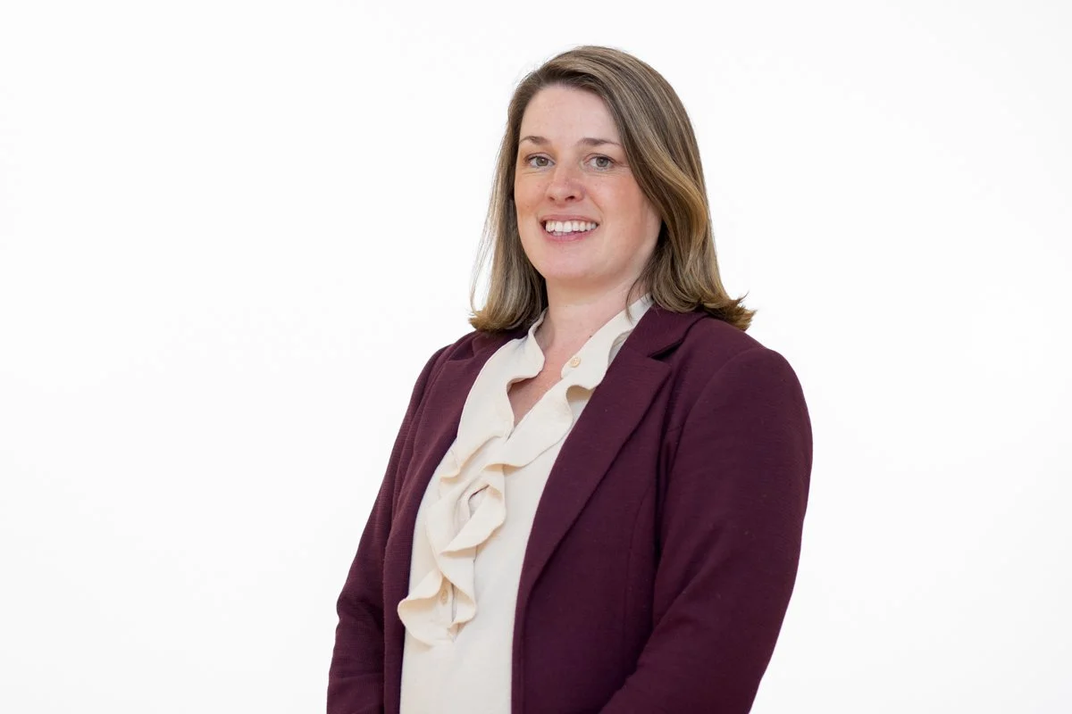 A woman with shoulder-length brown hair, wearing a white blouse with ruffles and a maroon blazer, smiling against a plain white background.
