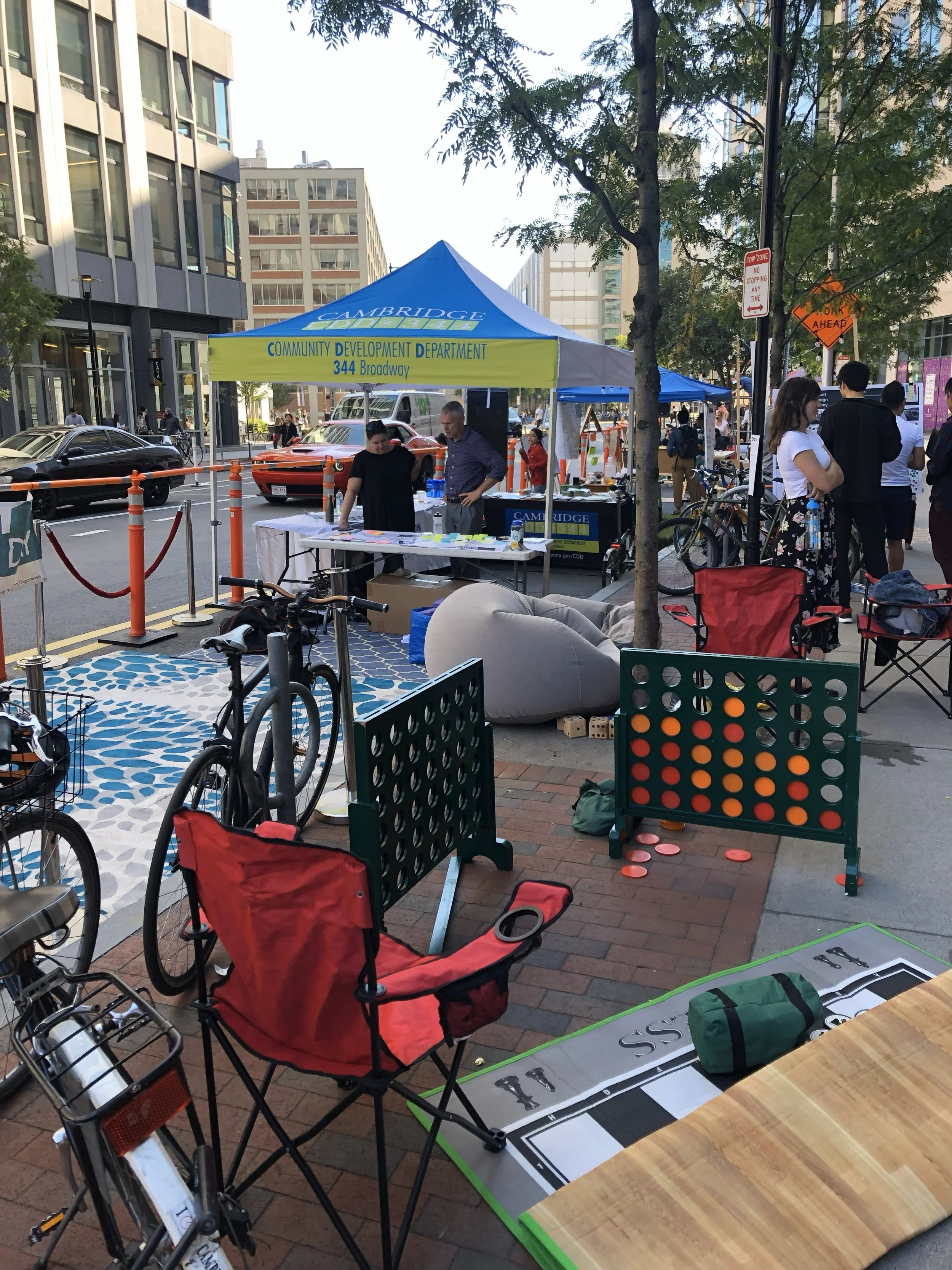 Outdoor street scene with a tent labeled 'Cambridge Community Development Department' and address '344 Broadway,' people standing and talking, bicycles parked, and various chairs and games set up on the sidewalk.