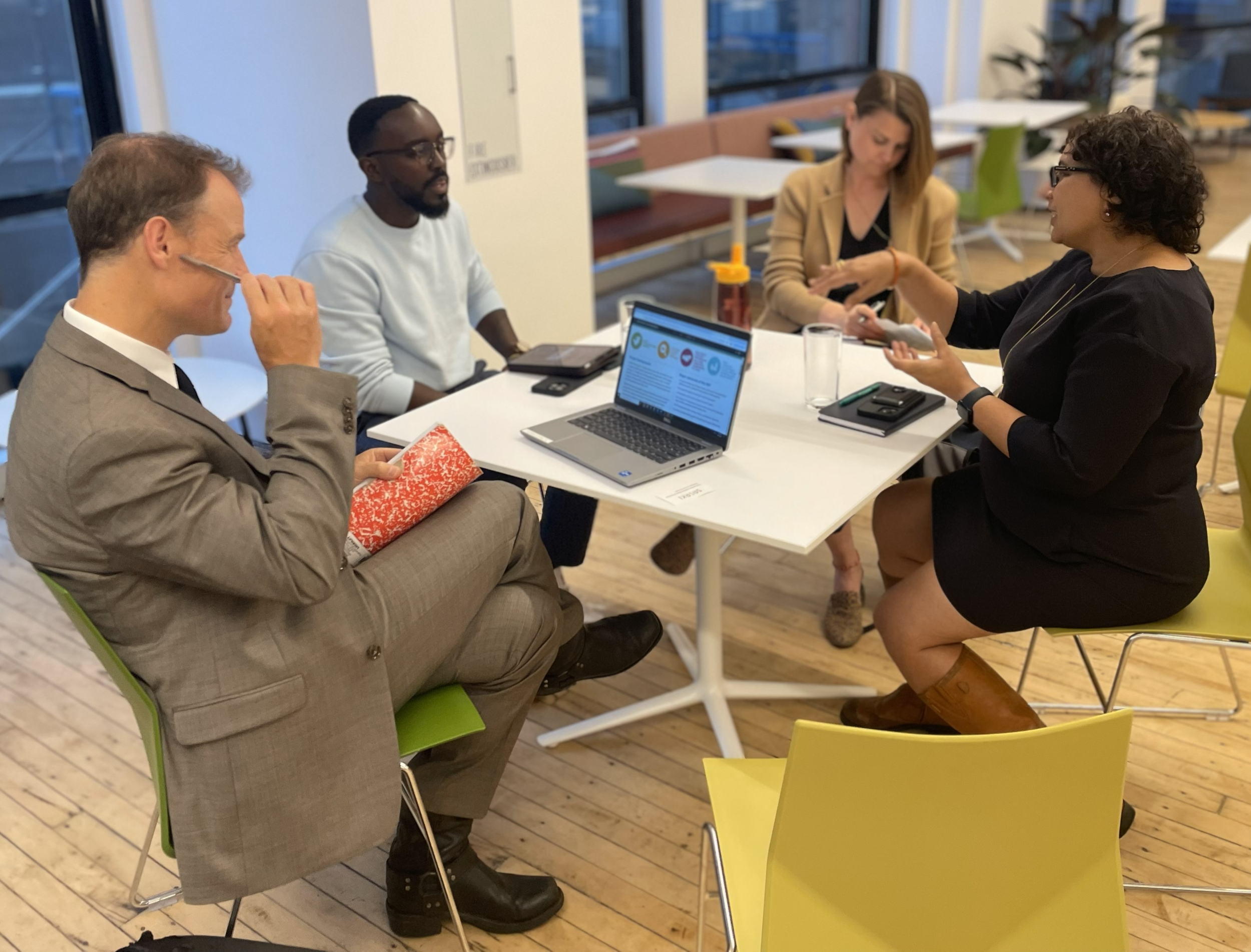 A group of four professionals collaborate before a speaking session in an office, sitting around a white table with laptops, notebooks, and drinks.
