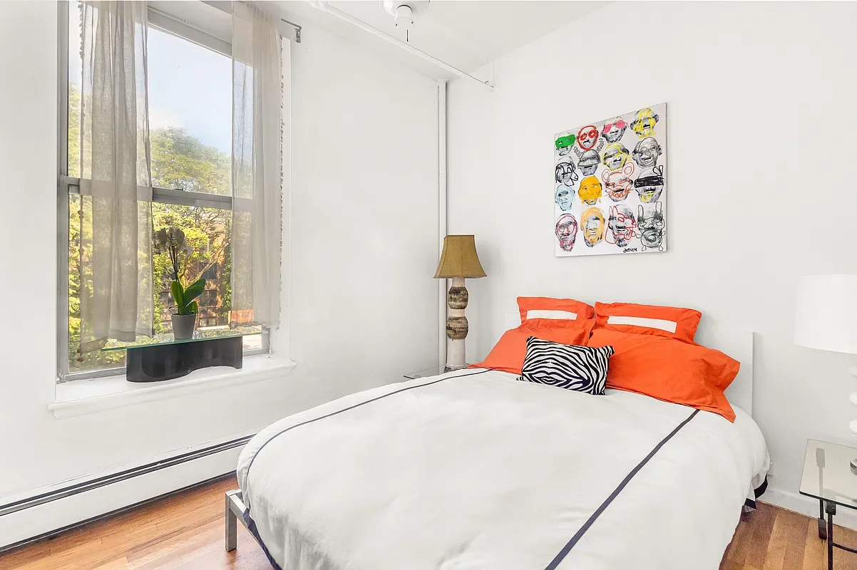 A bedroom with white walls and a large window with sheer curtains, a bed with orange pillows and a black and white zebra print pillow, a beige lamp with a textured base, a colorful artwork hanging above the bed, and a glass table on the side.