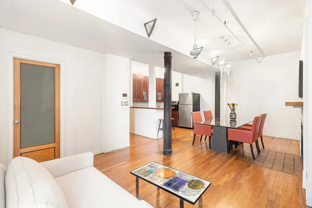 Living room and dining area with hardwood floors, white walls, a white sofa, a glass-top dining table with four pink chairs, a vase with flowers, a stainless steel refrigerator, and a kitchen area with wooden cabinets.
