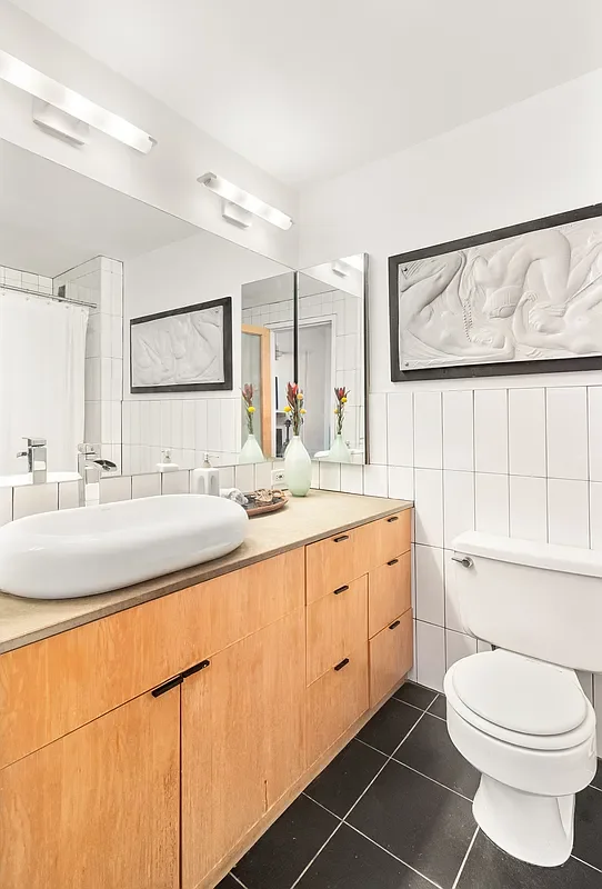 Modern bathroom with white tiled walls, black floor tiles, a wooden vanity with a vessel sink, a large mirror, and framed art.