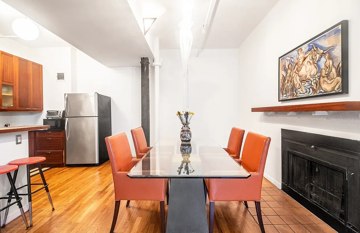 Dining room with a glass-top table, six orange chairs, a decorative vase in the center, wooden flooring, a painting on the wall, a fireplace, a refrigerator, and kitchen cabinets.