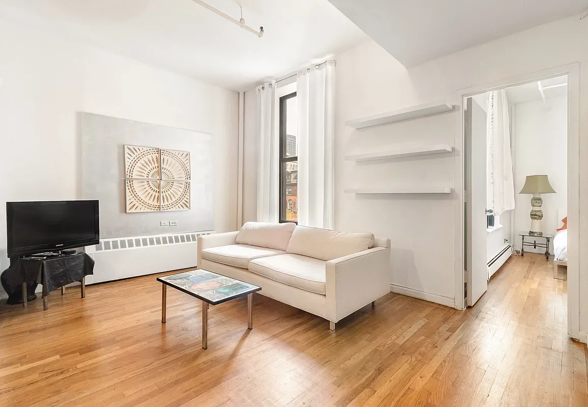 Living room with white walls, hardwood floor, white sofa, black TV on a small stand, coffee table with colorful top, three white wall shelves, large window with white curtains, doorway leading to bedroom with bed and nightstand.