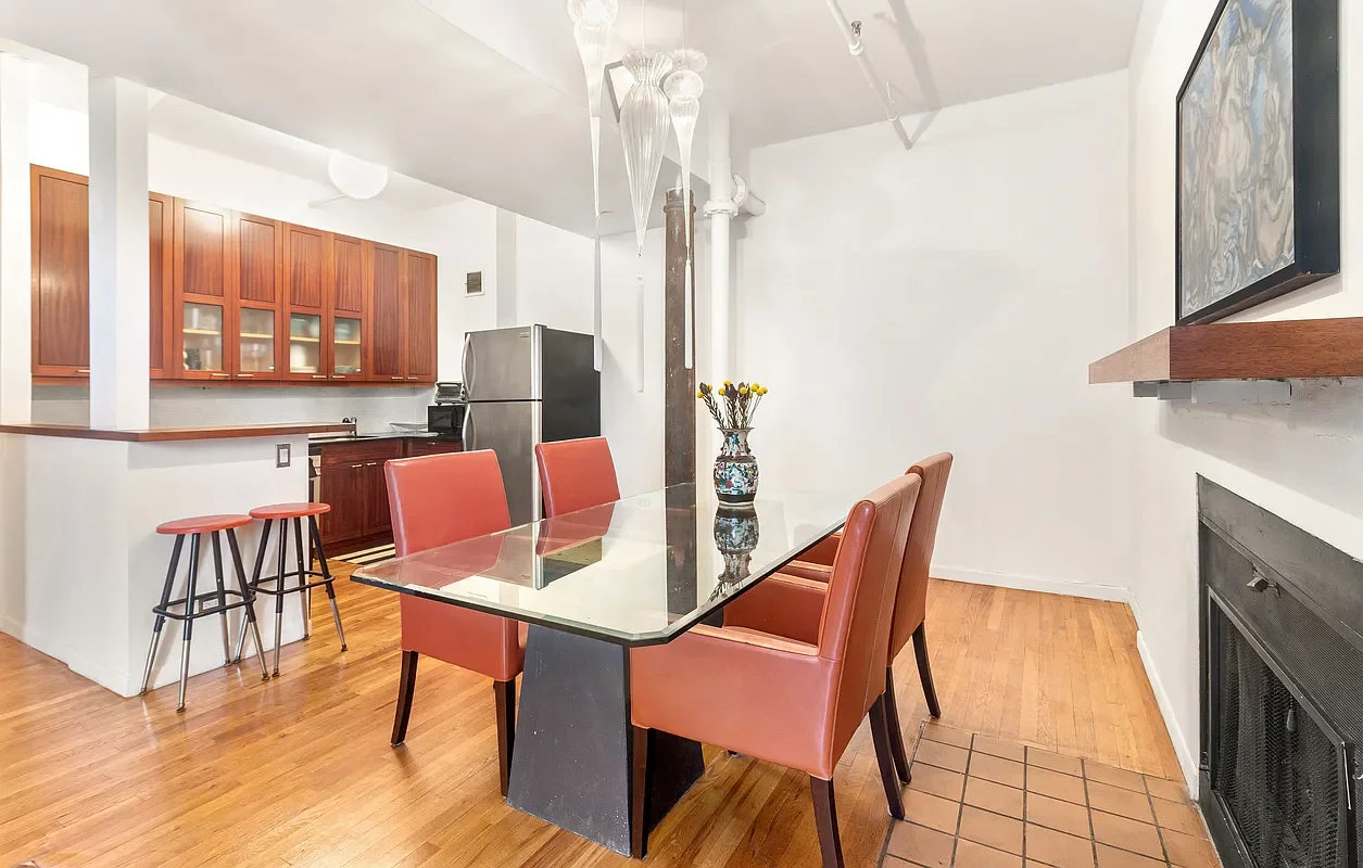 Dining room with a glass-top table, five orange chairs, a vase with yellow flowers, and a kitchen in the background with wooden cabinets and a stainless steel refrigerator.