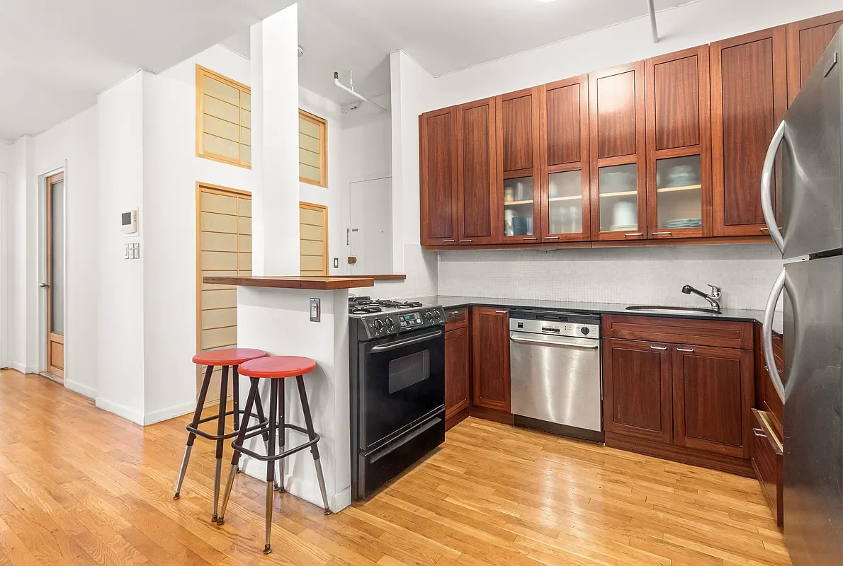 Kitchen with wooden cabinets, black stove, stainless steel dishwasher, stainless steel refrigerator, small counter space, two red bar stools, hardwood floors.