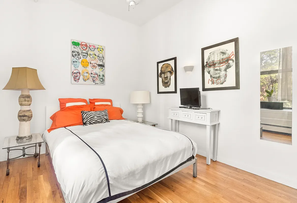 A bedroom with a bed, orange pillows, black and white zebra print pillow, a large beige lamp, a white nightstand, a television on a white console, and framed artwork on the wall.