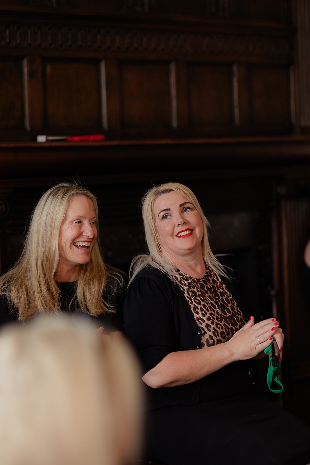 Karen Weaver and Adrea Peters sit together, one with blonde hair laughing and the other with blonde hair wearing a leopard print top, smiling and holding green and red items, in a wood-paneled room.