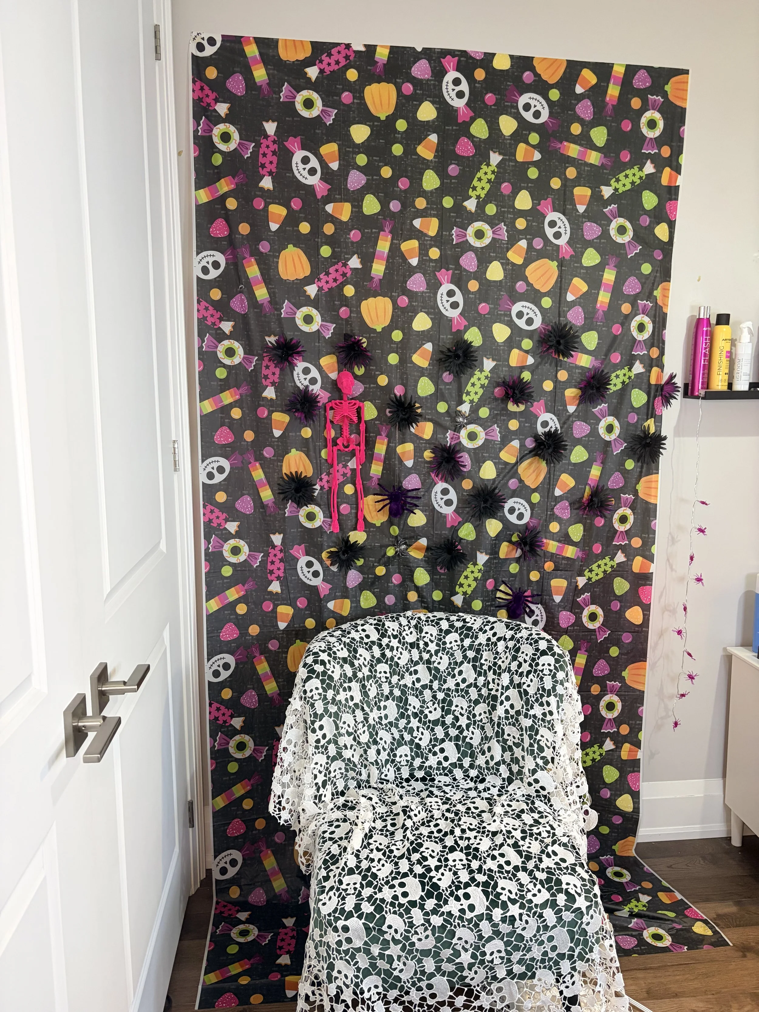 A room with a colorful Halloween-themed backdrop featuring skulls, pumpkins, candies, and eyeballs, with a white lace-covered chair in front.