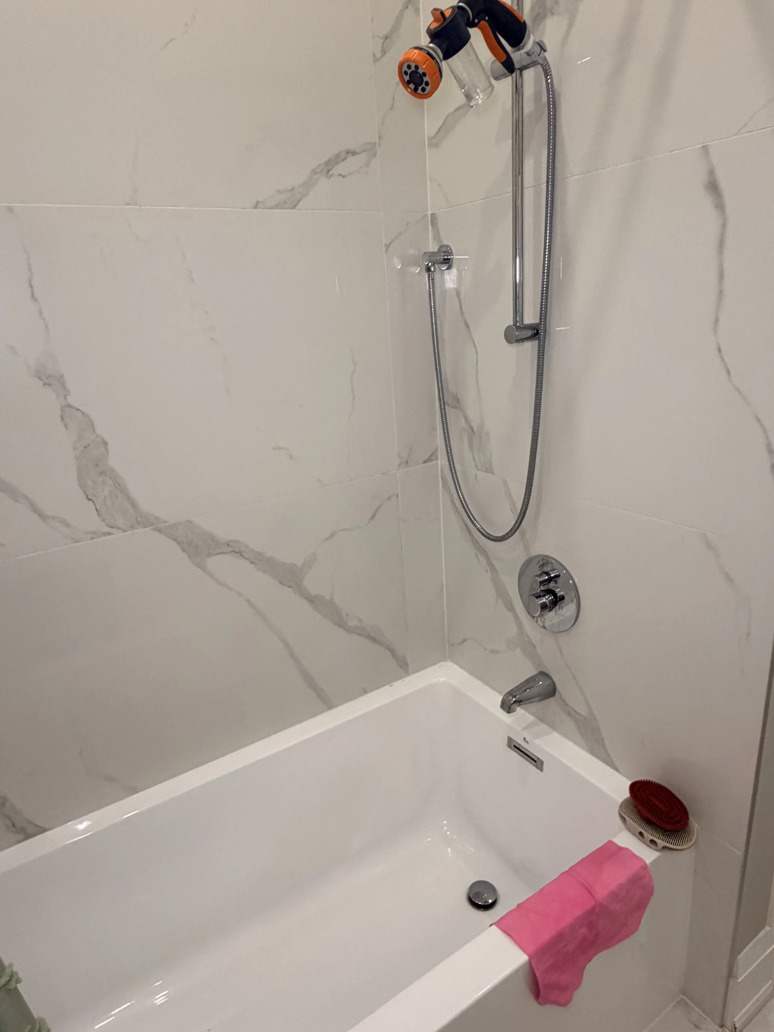Bathroom bathtub with marble tiled walls, a handheld shower, a drain, a faucet, a pink towel hanging over the edge of the tub, a red scrub brush, and a pink cloth near the drain.