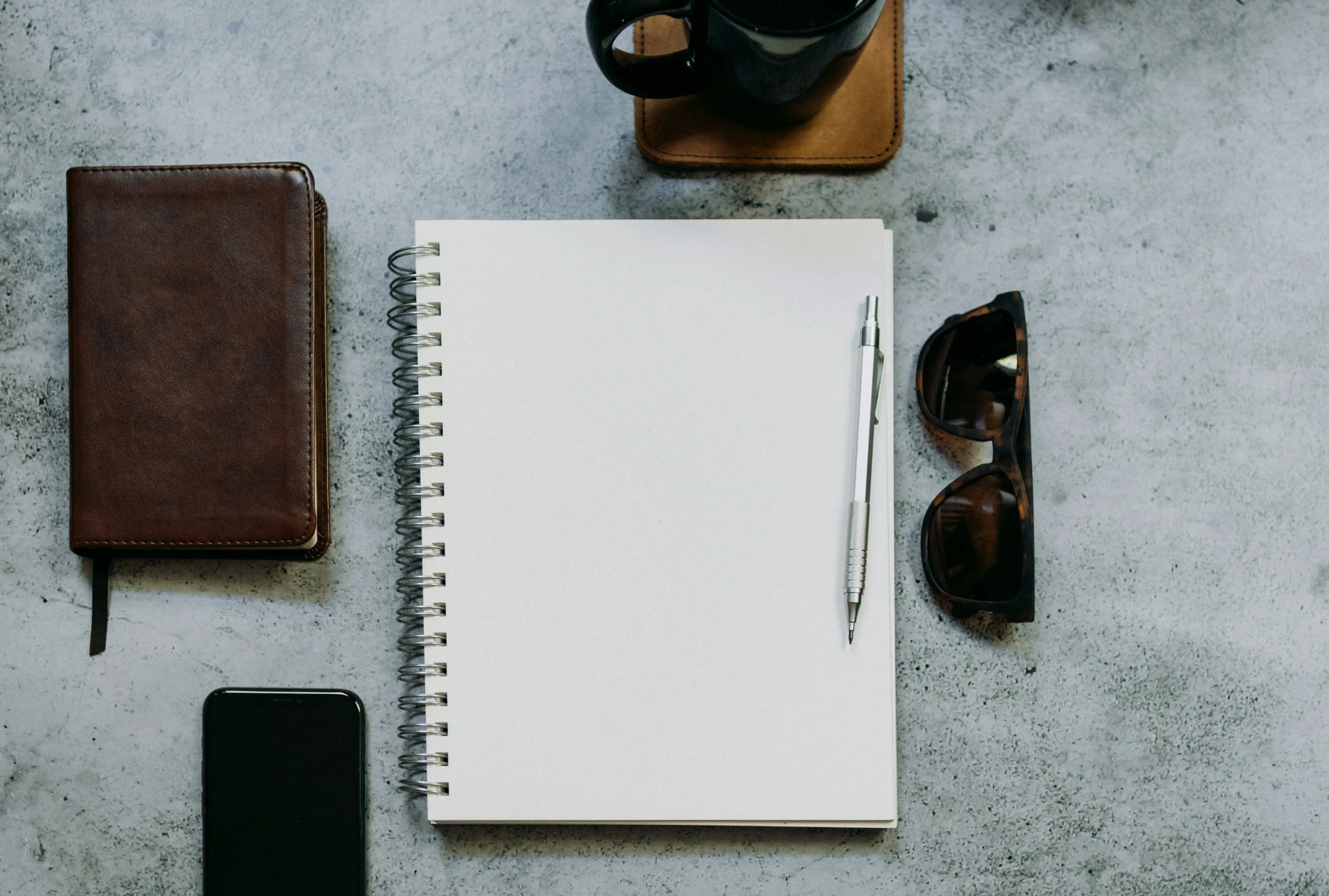 Flat lay of a workspace on a gray concrete surface, including a brown leather wallet, a black smartphone, a white spiral notebook with a silver pen, a black coffee mug on a brown leather coaster, and a pair of tortoiseshell sunglasses.
