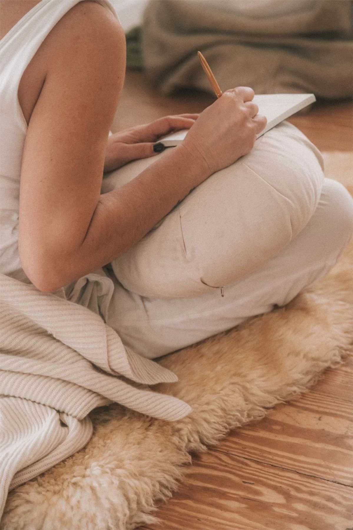 A person sitting on a beige shaggy rug on a wooden floor, writing in a notebook with a pen, wearing a sleeveless top and light-colored pants.