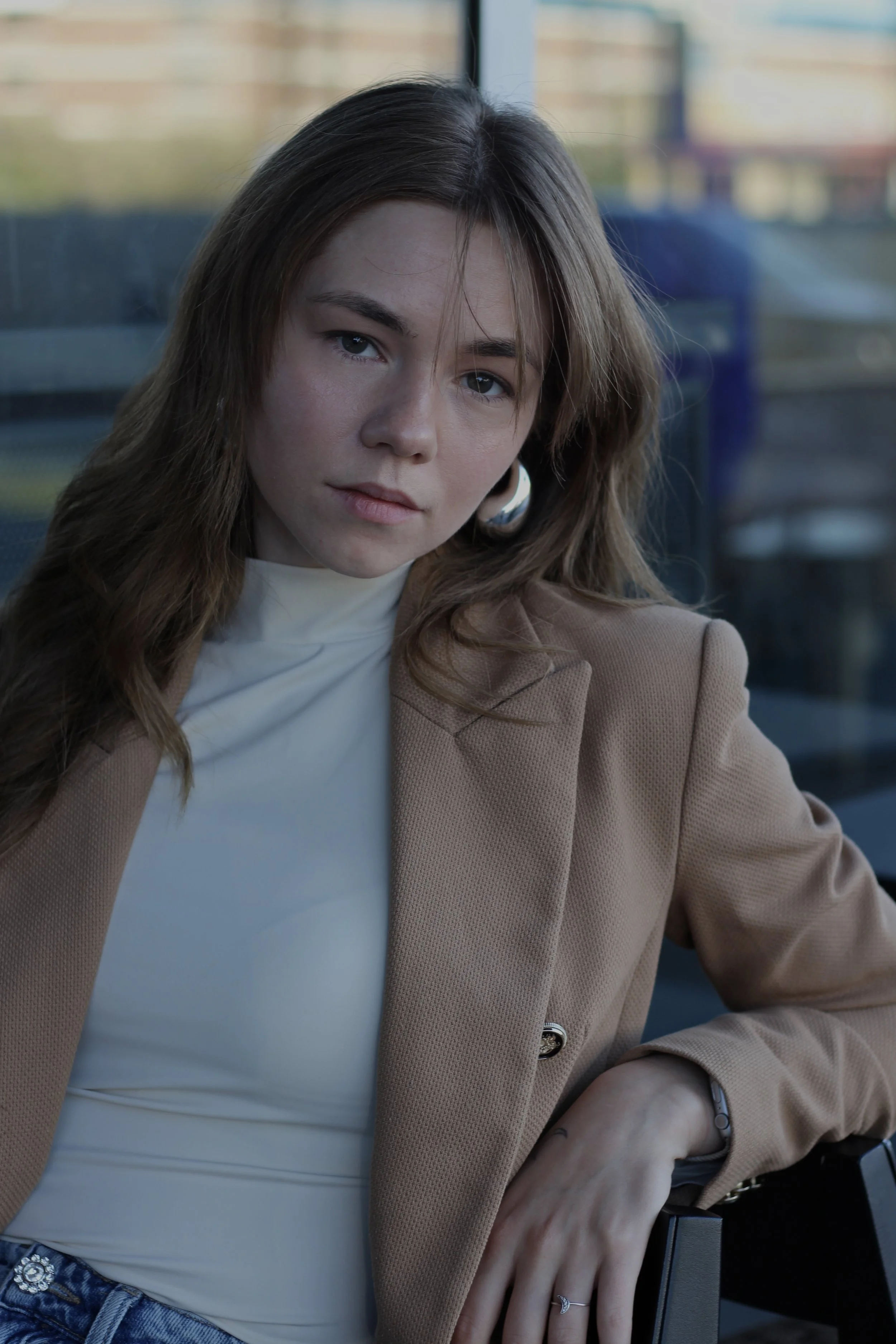 A young woman with brown hair and large hoop earrings sitting near a window, wearing a beige blazer over a white turtleneck, and looking directly at the camera.