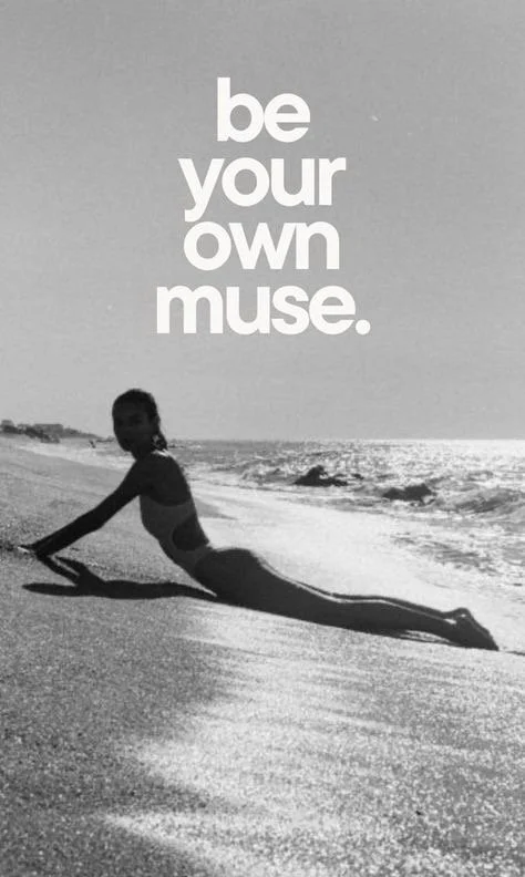 A person lying on the beach with their body stretched out, creating the illusion of a mermaid tail, with the ocean in the background. The text reads 'be your own muse.'