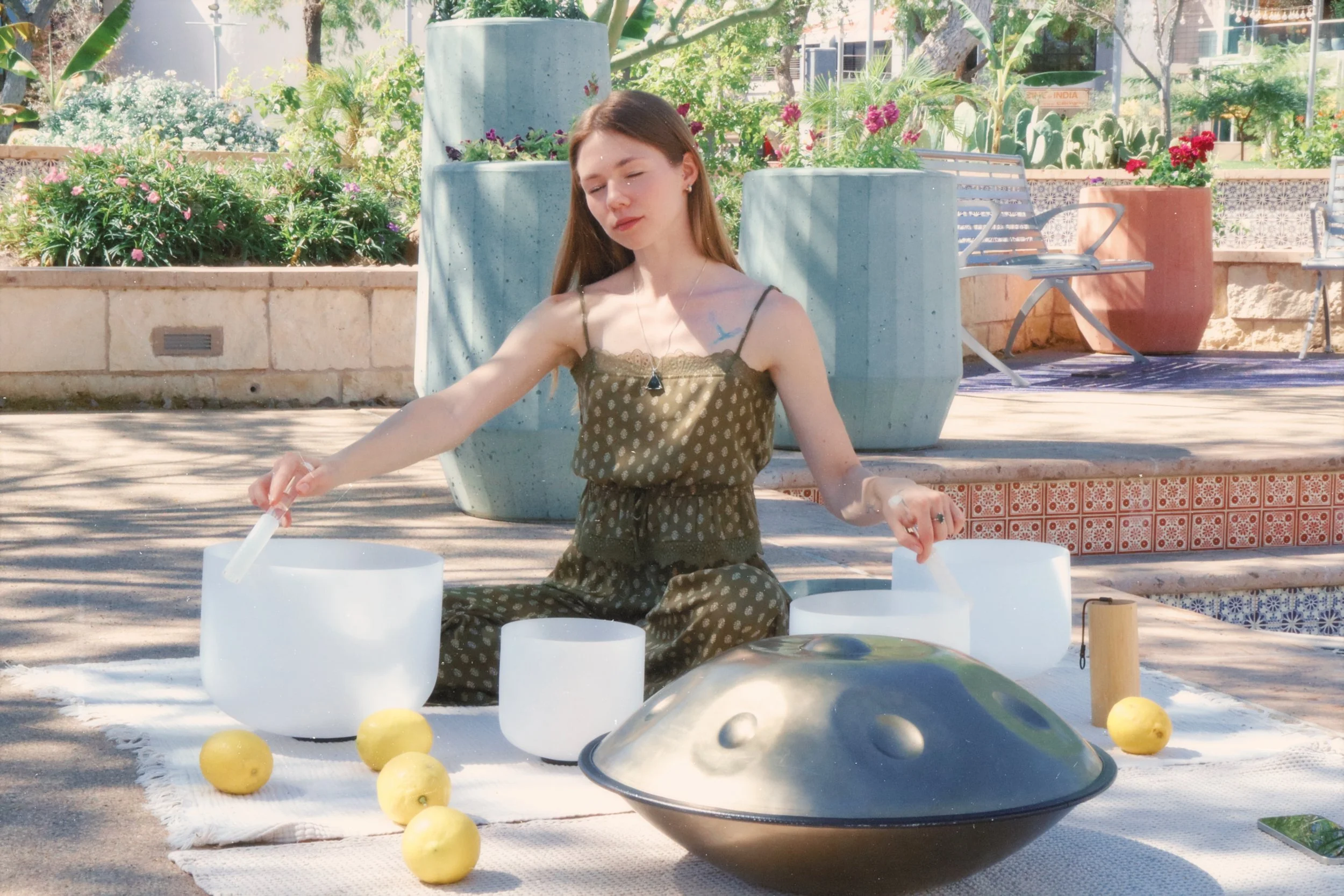 A young woman performing a singing bowl meditation outdoors surrounded by lemons, large ceramic planters, and lush greenery.