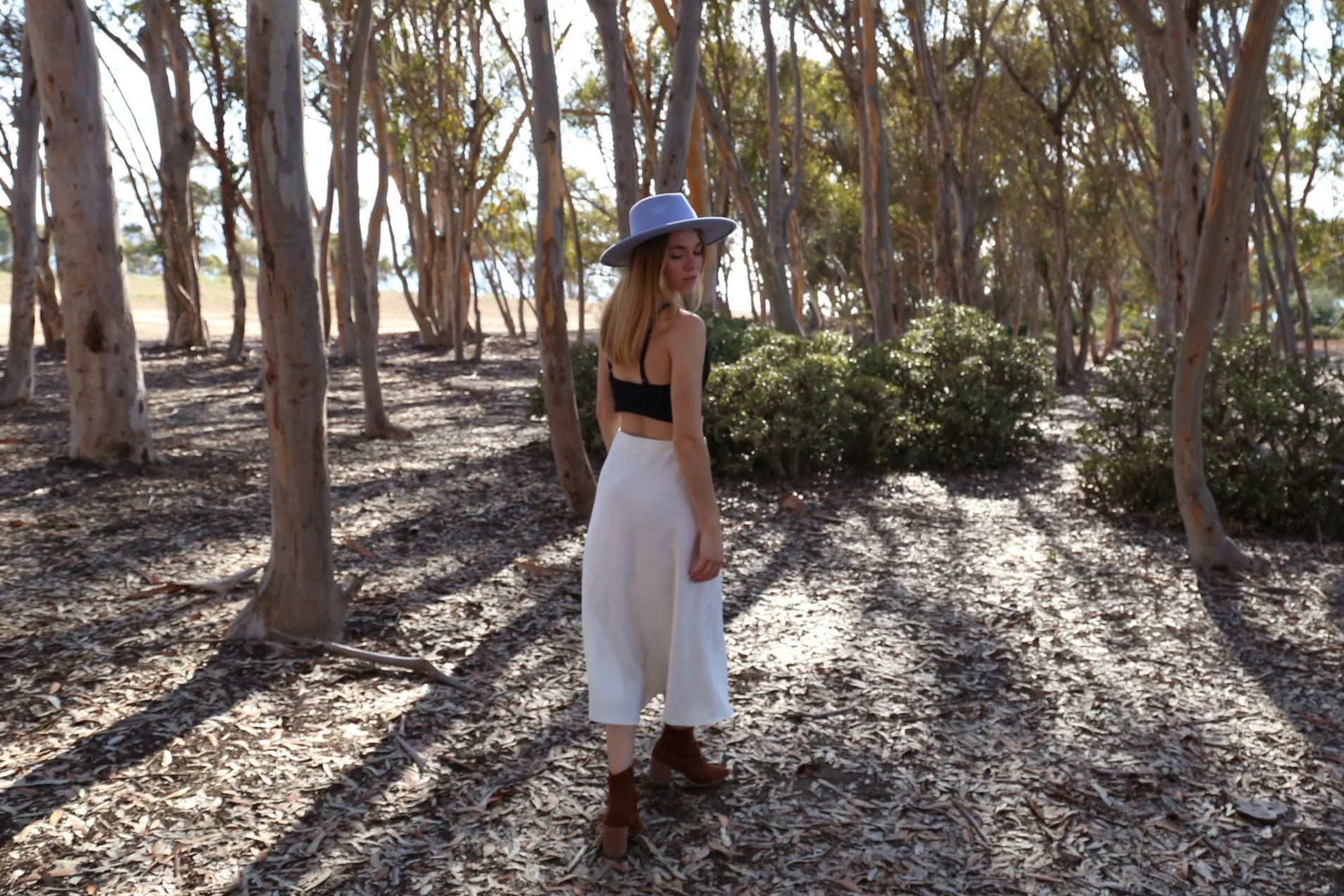 A woman in a black crop top, white pants, brown ankle boots, and a wide-brimmed blue hat standing in a forest with tall trees and leaf-covered ground.