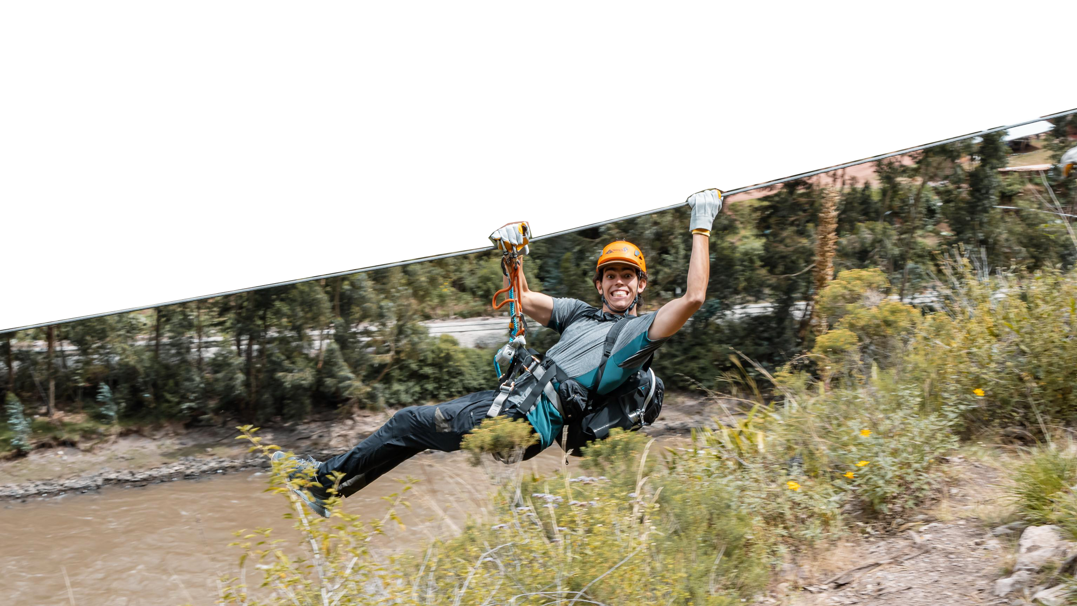Young man ziplining over a river, wearing a helmet, harness, and gloves, smiling as he holds onto the zipline cable in an outdoor wooded area.