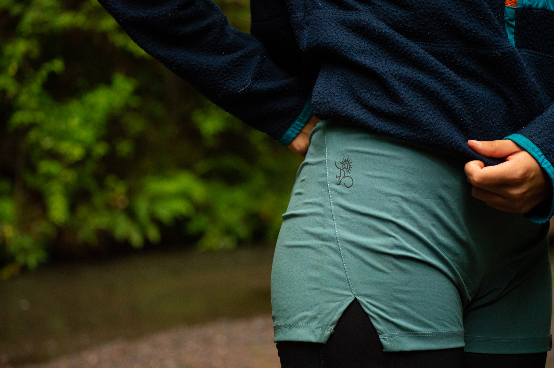 Close-up of a person wearing green outdoor shorts with a small logo near the waistband, and a navy fleece jacket with teal accents, standing in a natural setting with greenery in the background.