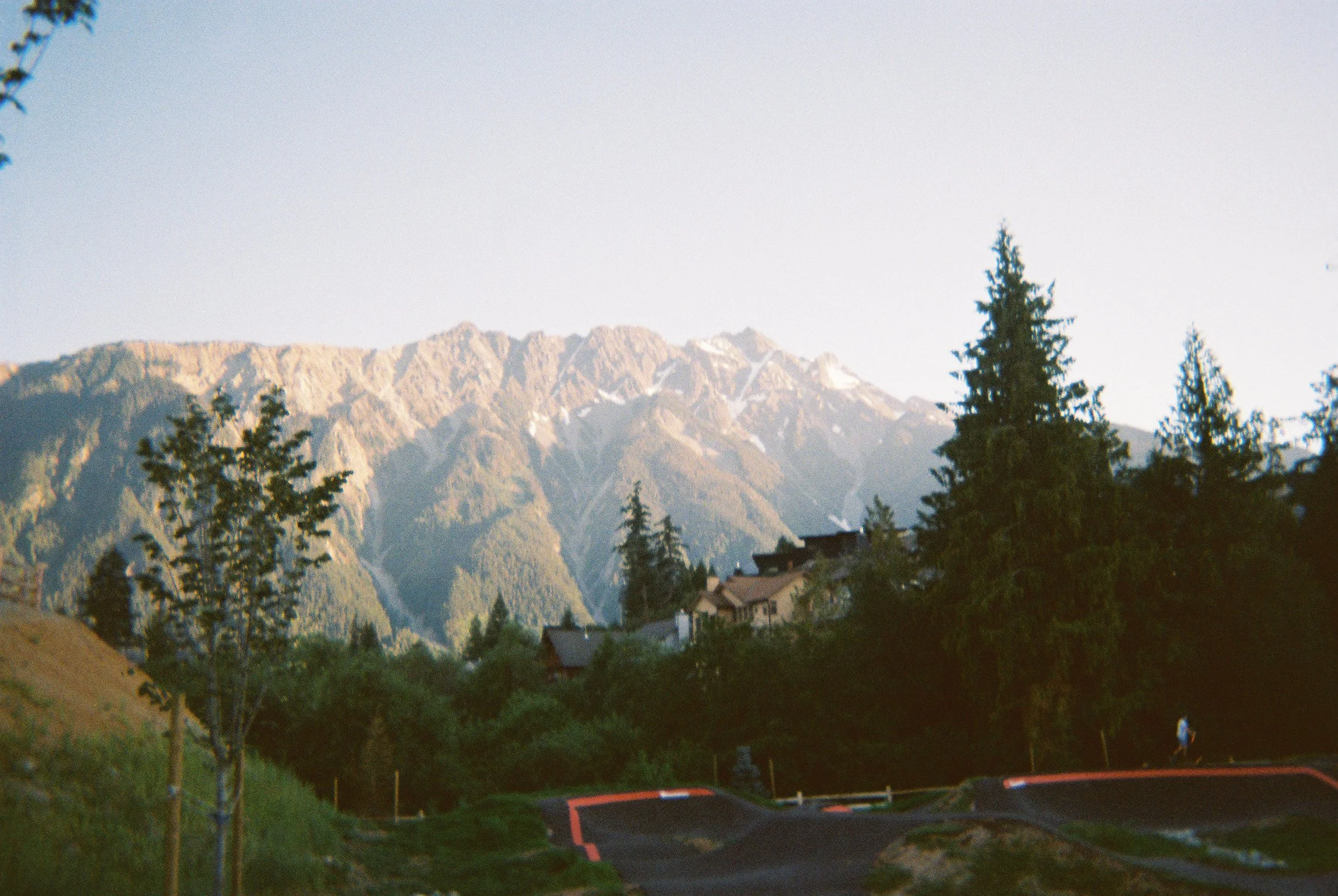 Mountains with snow patches, trees, and houses on a hillside at sunset.