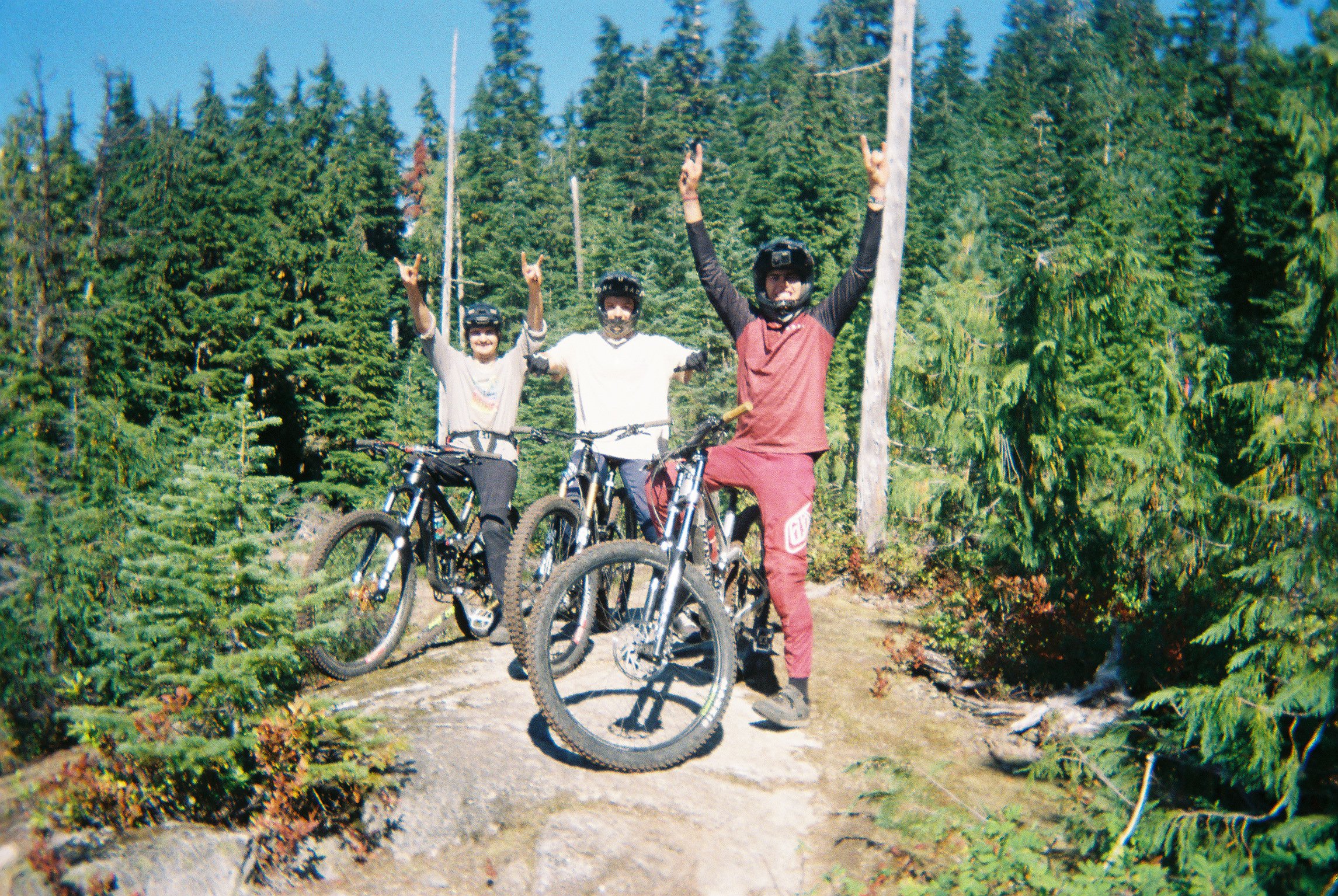 Three people on mountain bikes celebrating with raised hands in a forested outdoor area under a clear blue sky.