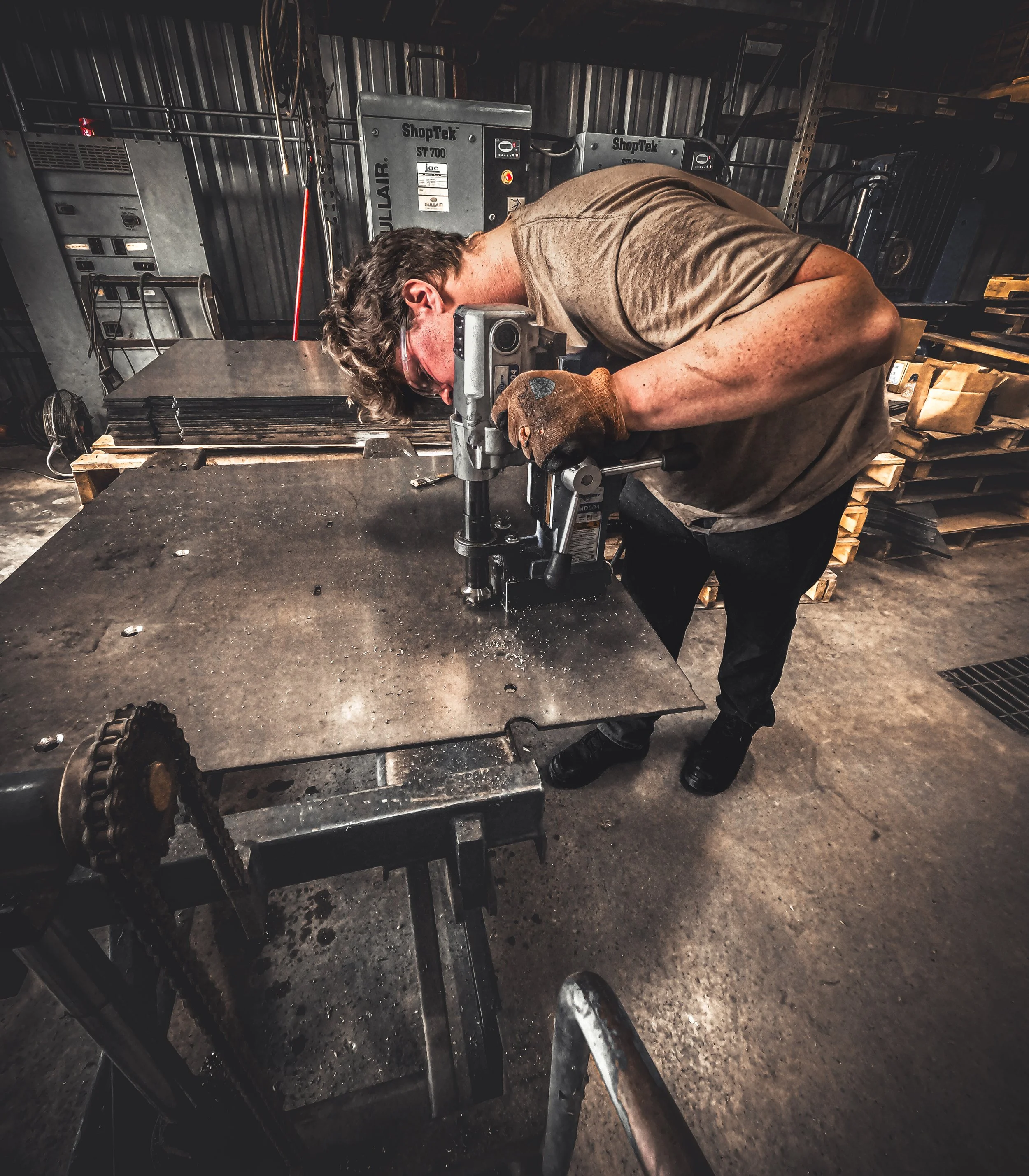 Man wearing safety glasses and gloves using a power drill on a metal workpiece in a workshop.