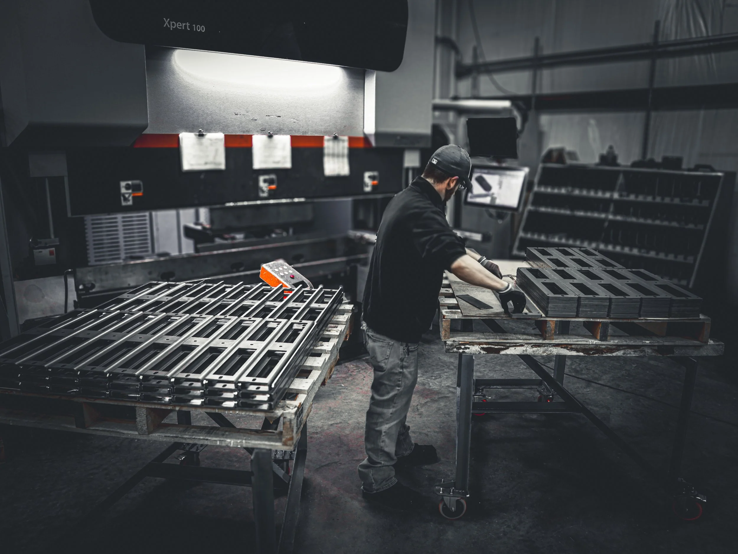 A worker wearing a baseball cap, glasses, and gloves working on metal components at a manufacturing station in an industrial factory, with machinery and equipment in the background.