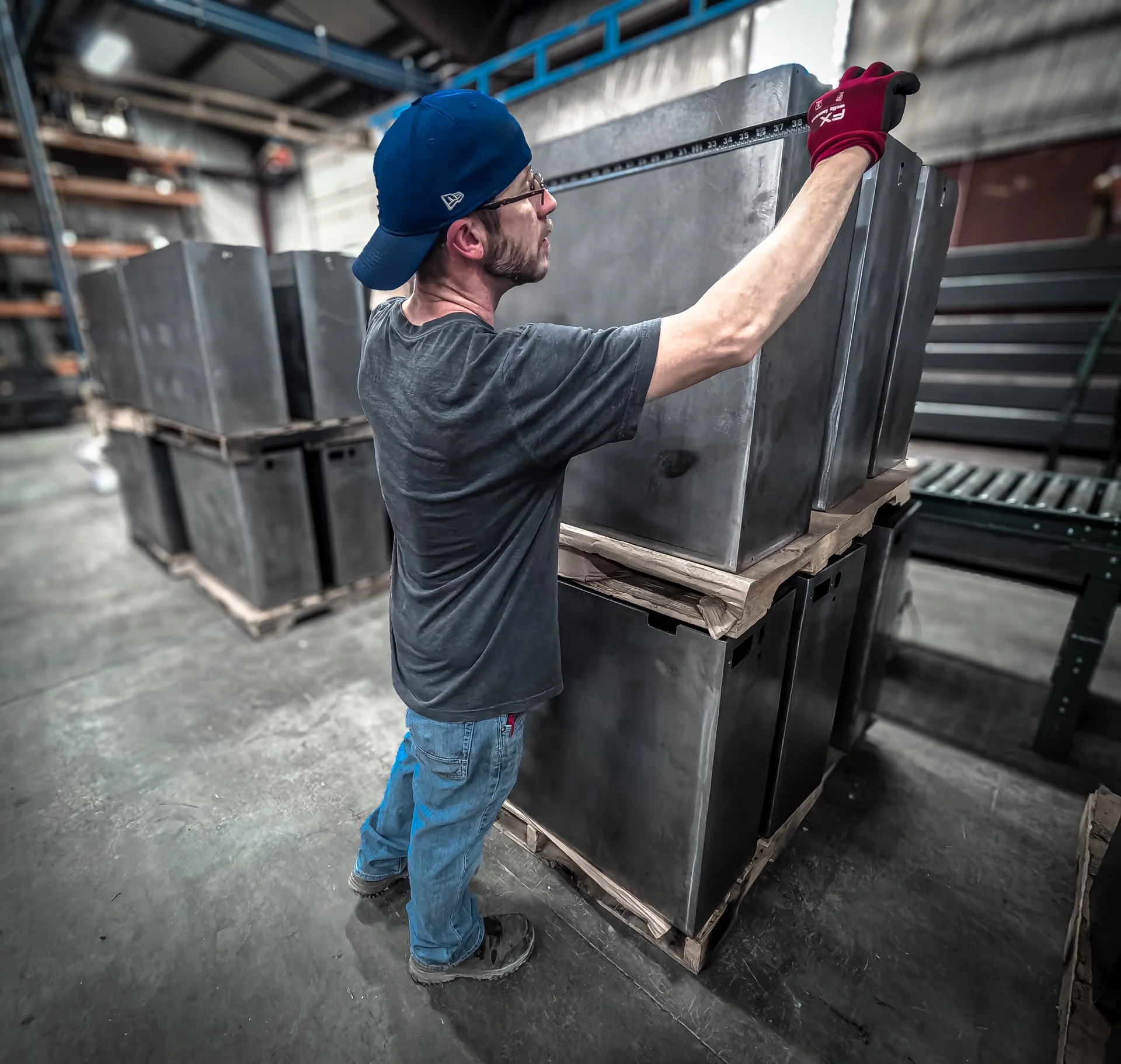 A man wearing a blue cap, glasses, a dark t-shirt, and jeans measuring a large metal box with a tape measure in an industrial warehouse.