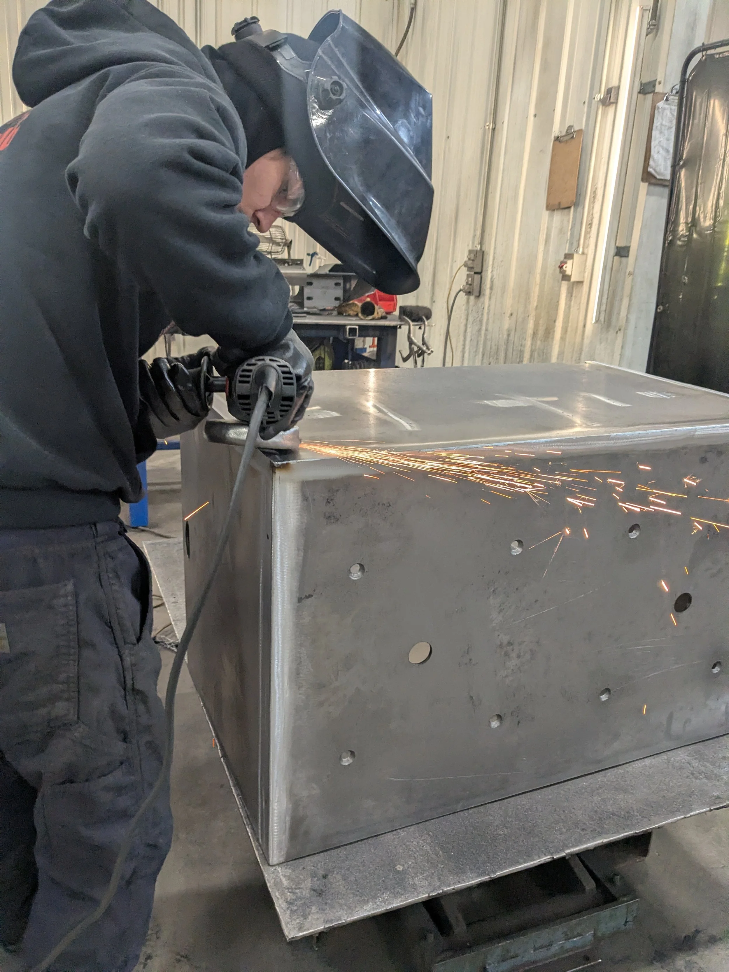 A person grinding on a large metal box in a workshop, wearing protective gear including a welding helmet and gloves.