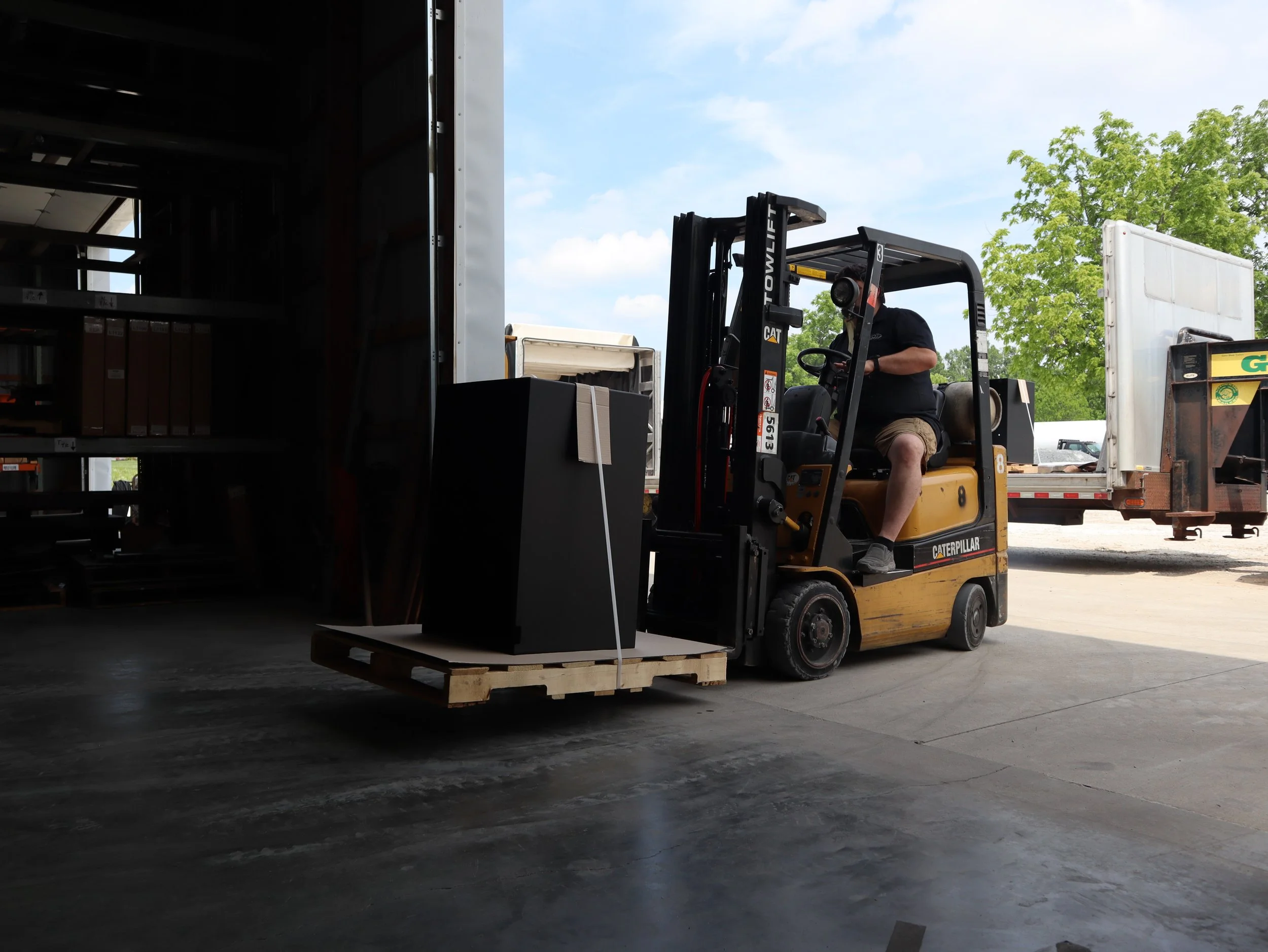 A man operating a yellow Caterpillar forklift with black accents is moving a wooden pallet with a large black electronic device into a storage container. The forklift is outside on concrete, beside a large open storage area, with trees and a blue sky in the background.