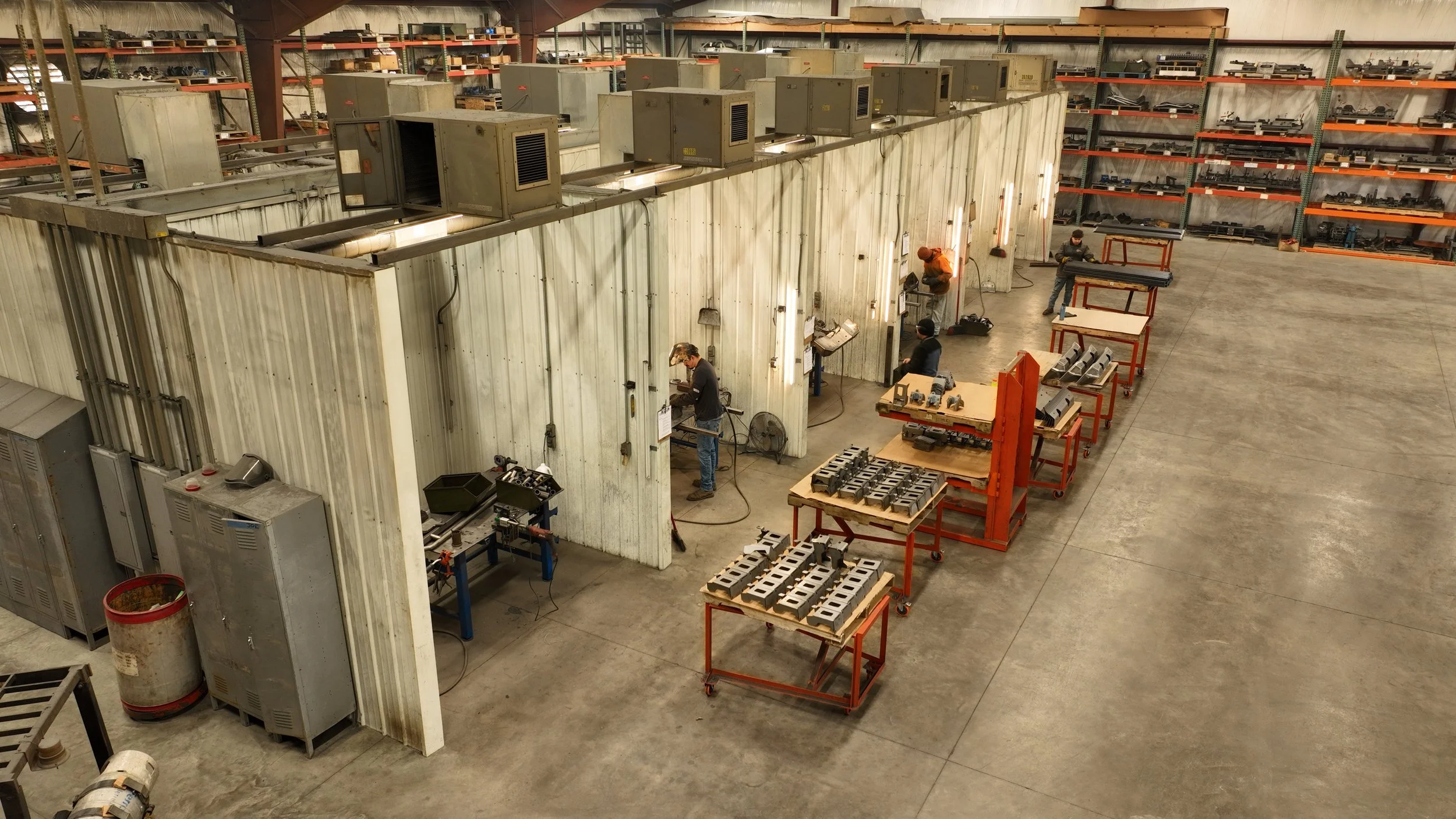 Workers welding metal parts inside a large industrial facility with storage shelves filled with various equipment in the background. Metal workbenches with tools and metal components are arranged on carts.