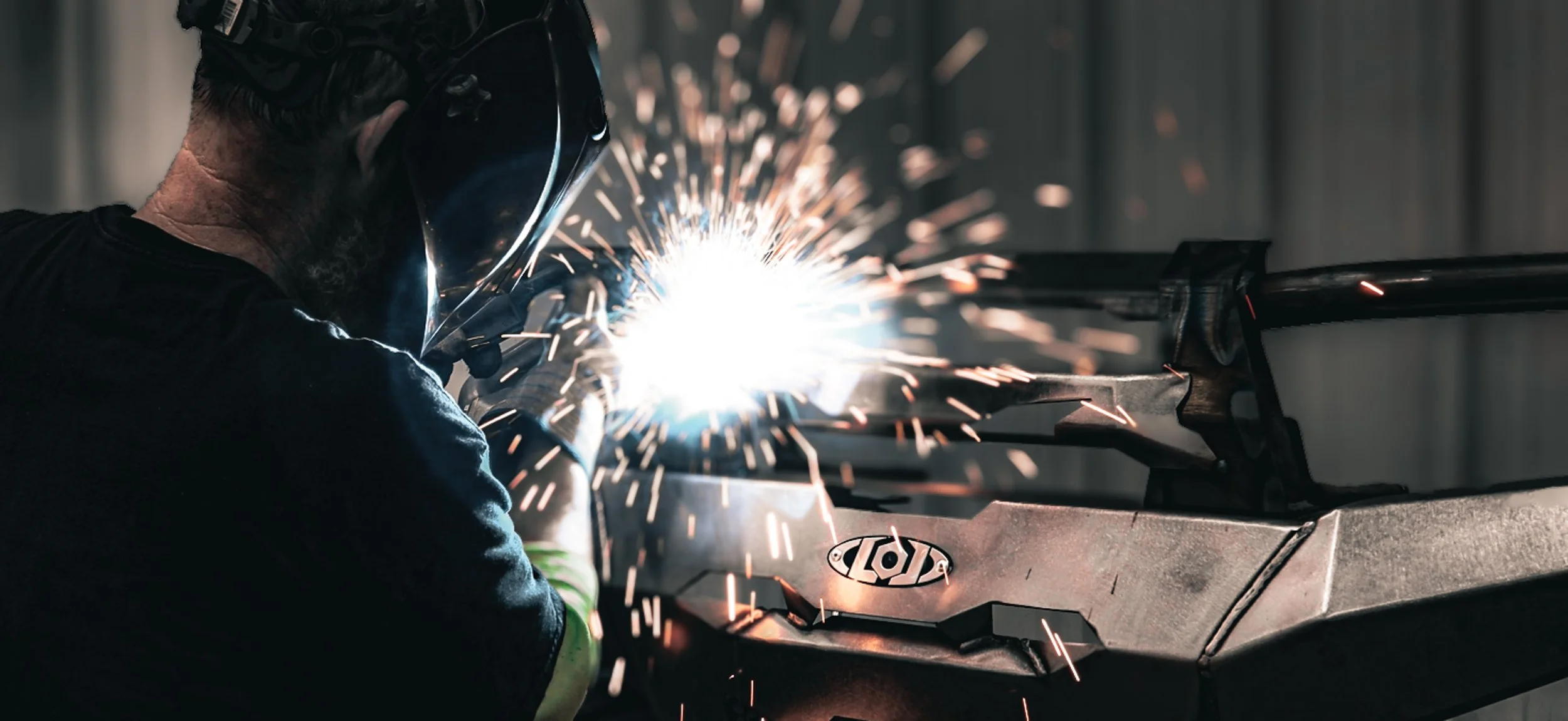 A person welding metal, with sparks flying, wearing protective gear including a helmet and gloves, working on a metal frame.