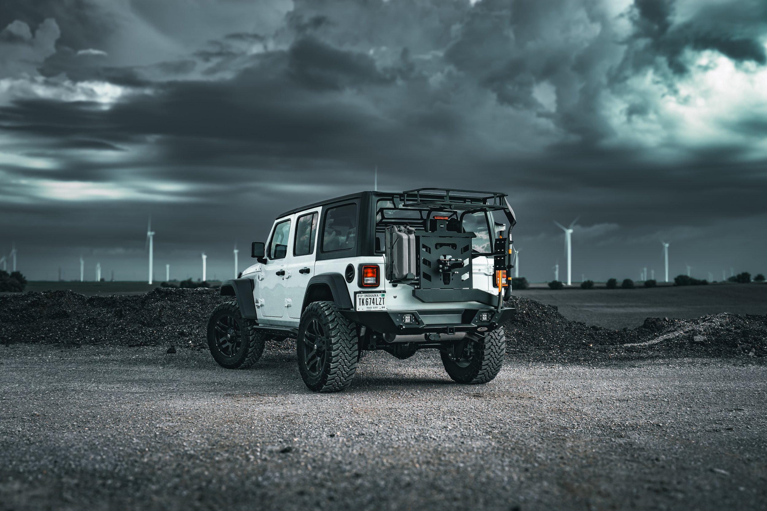 A rugged off-road vehicle with a black and white color scheme, large tires, and mounted equipment on the back, parked on dirt beneath a dark, cloudy sky with wind turbines in the distance.