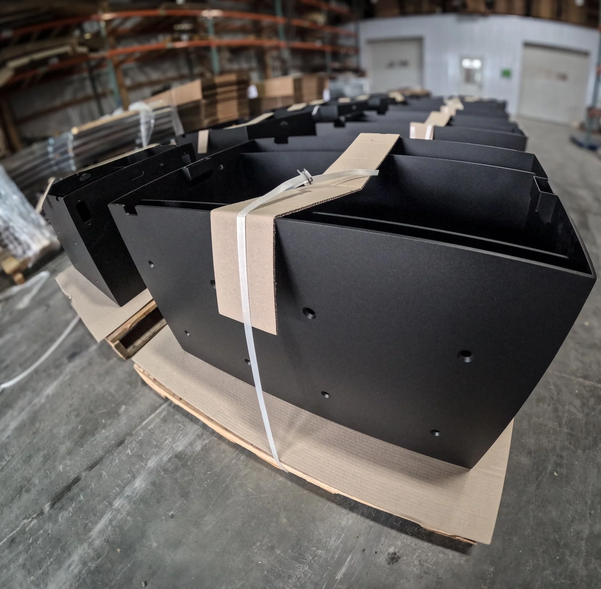 Black metal or plastic component secured with a beige strap on a cardboard pallet in a warehouse.