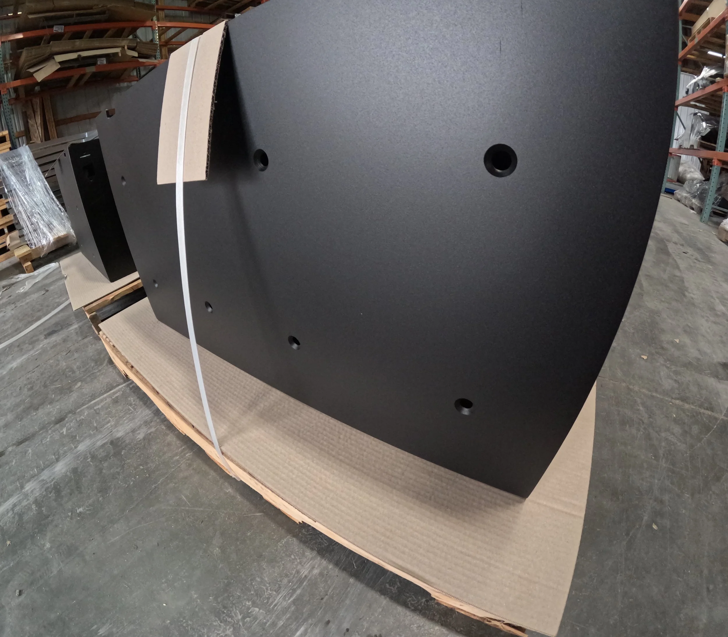 Large black speaker cabinet resting on a cardboard pallet in a warehouse.